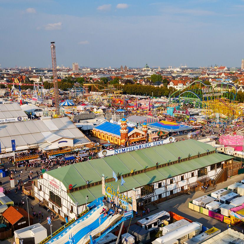 Luftaufnahme eines lebhaften Volksfestes mit bunten Fahrgeschäften, Zelten und Buden vor der Skyline einer Stadt bei klarem Himmel. Menschenmengen genießen Unterhaltung und traditionelle Feststimmung.