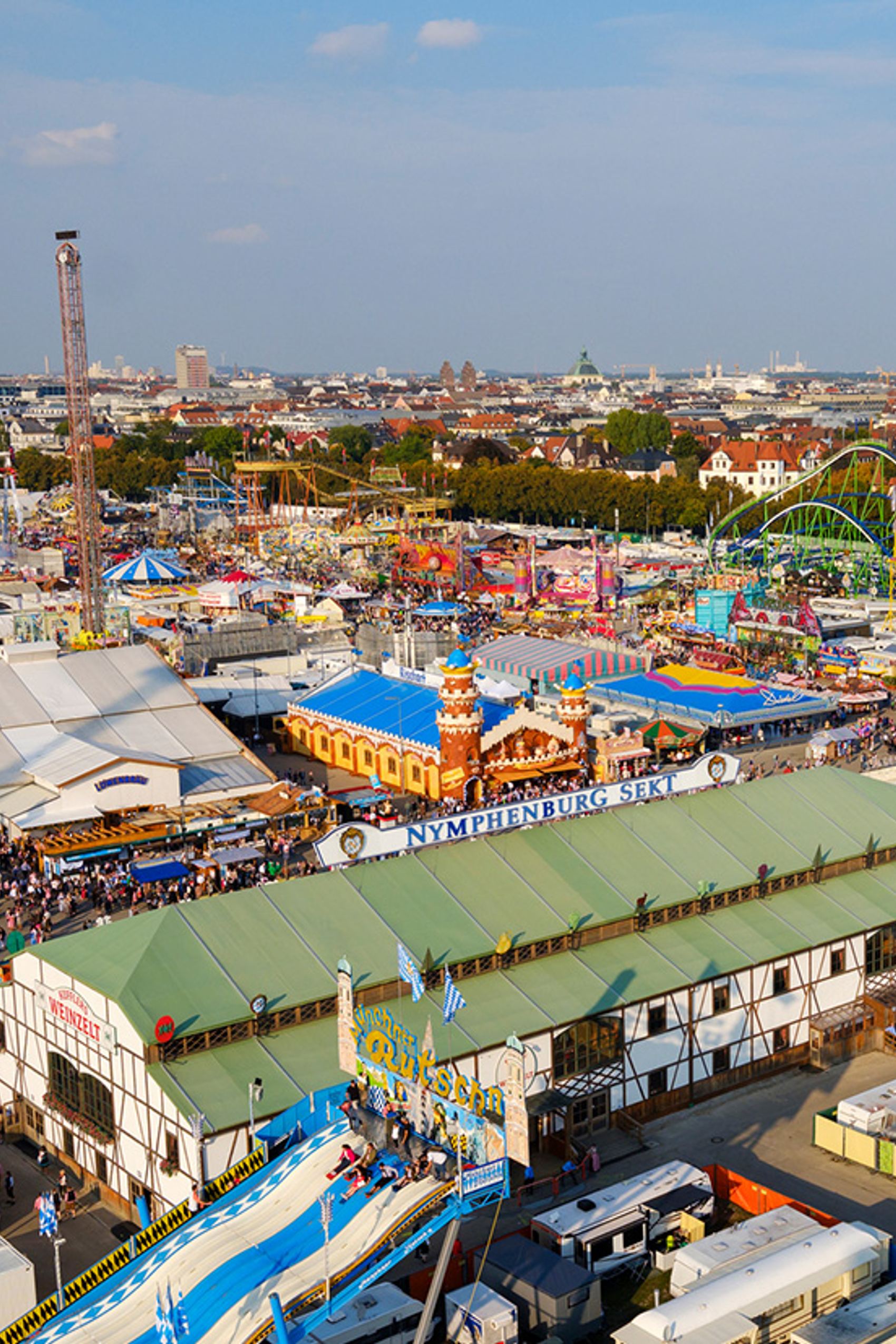 Luftaufnahme eines lebhaften Volksfestes mit bunten Fahrgeschäften, Zelten und Buden vor der Skyline einer Stadt bei klarem Himmel. Menschenmengen genießen Unterhaltung und traditionelle Feststimmung.