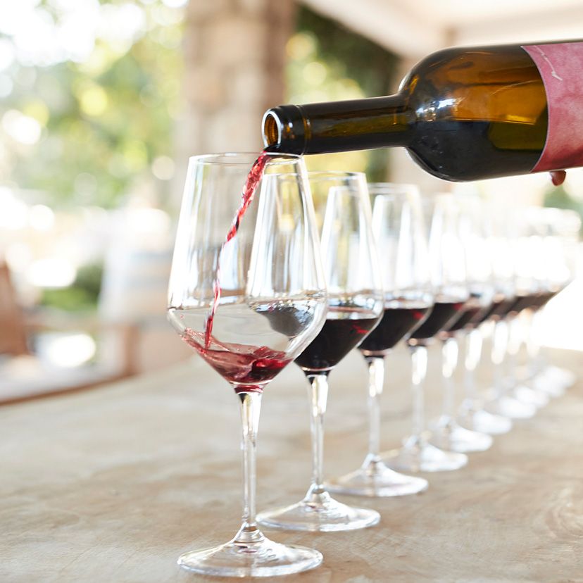 Roter Wein wird aus einer Flasche in ein Glas eingeschenkt, mehrere weitere halb gefüllte Weingläser stehen in einer Reihe auf einem hellen Tisch.