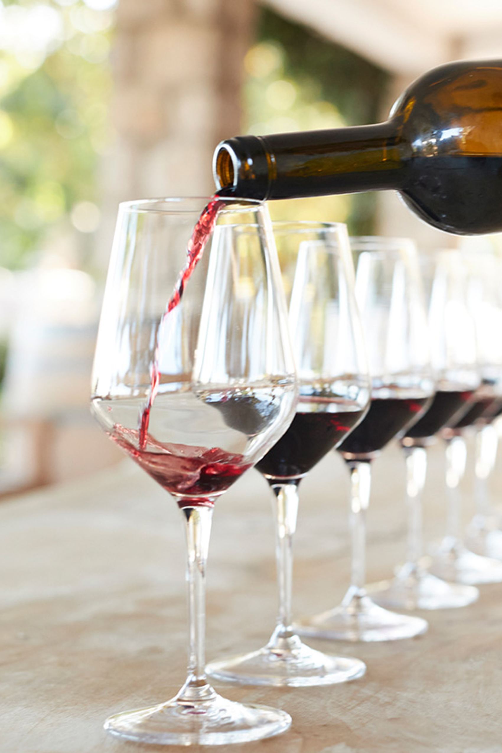 Roter Wein wird aus einer Flasche in ein Glas eingeschenkt, mehrere weitere halb gefüllte Weingläser stehen in einer Reihe auf einem hellen Tisch.