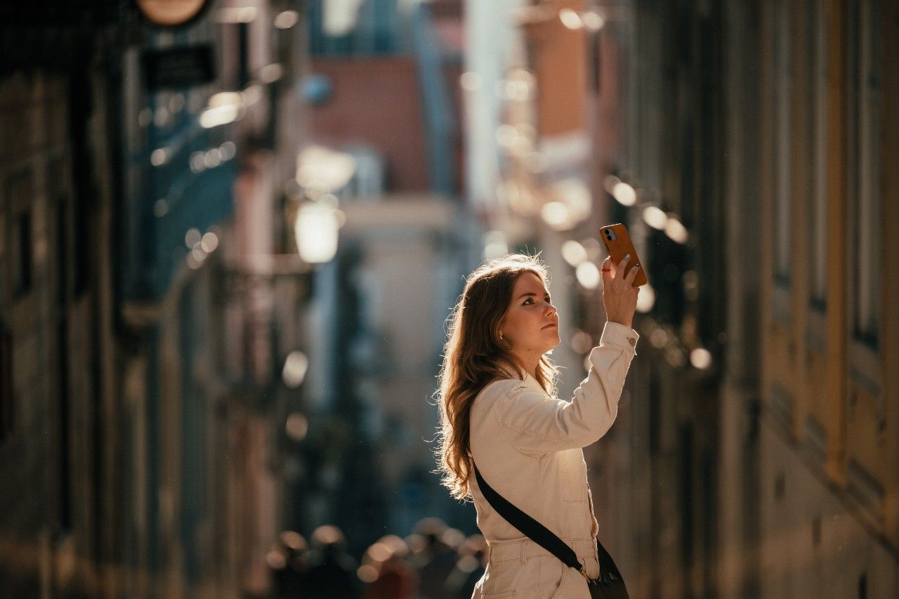 Junge Frau steht in einer sonnendurchfluteten Gasse und hält ihr Smartphone zum Fotografieren nach oben, wirkt konzentriert und neugierig.