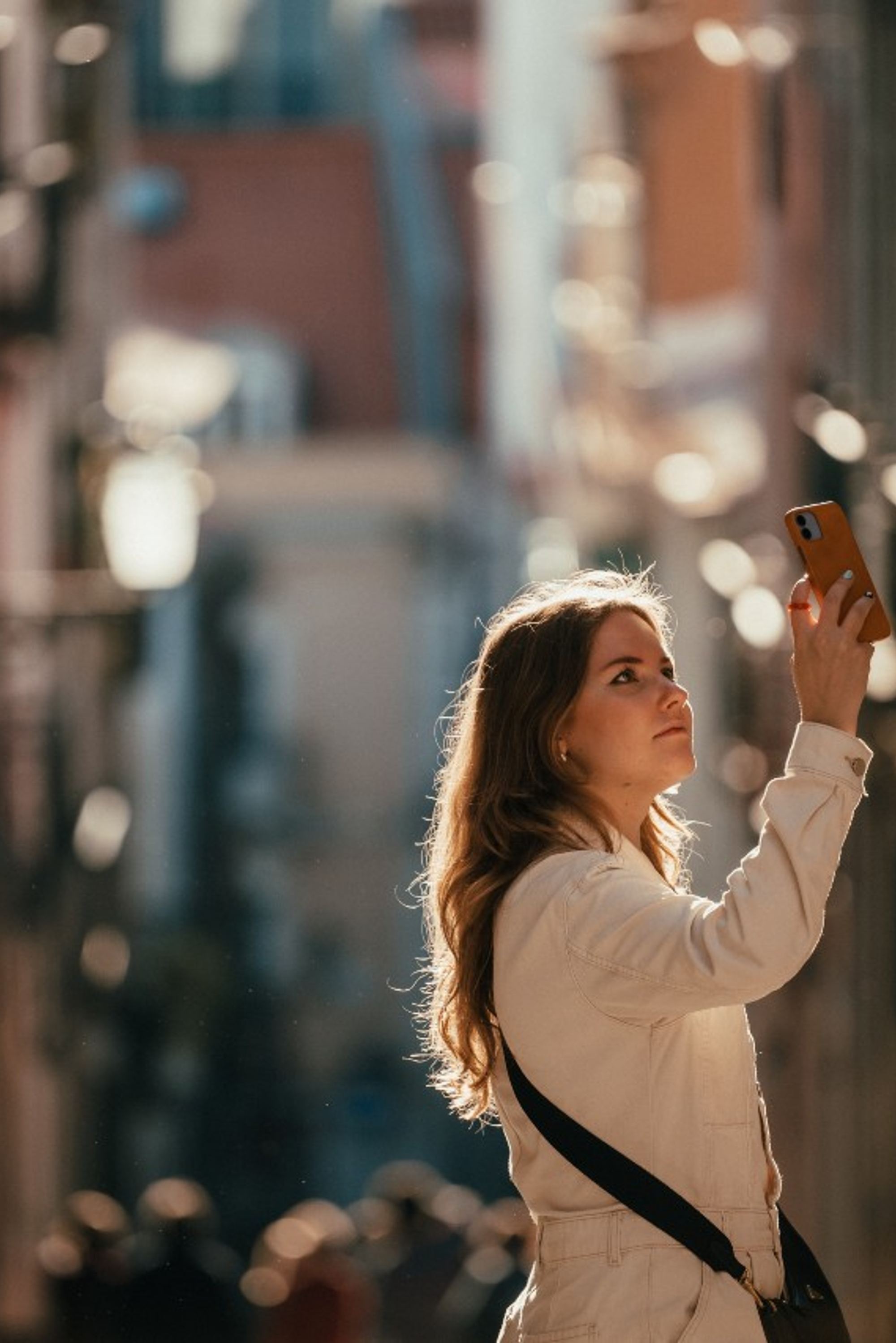 Junge Frau steht in einer sonnendurchfluteten Gasse und hält ihr Smartphone zum Fotografieren nach oben, wirkt konzentriert und neugierig.