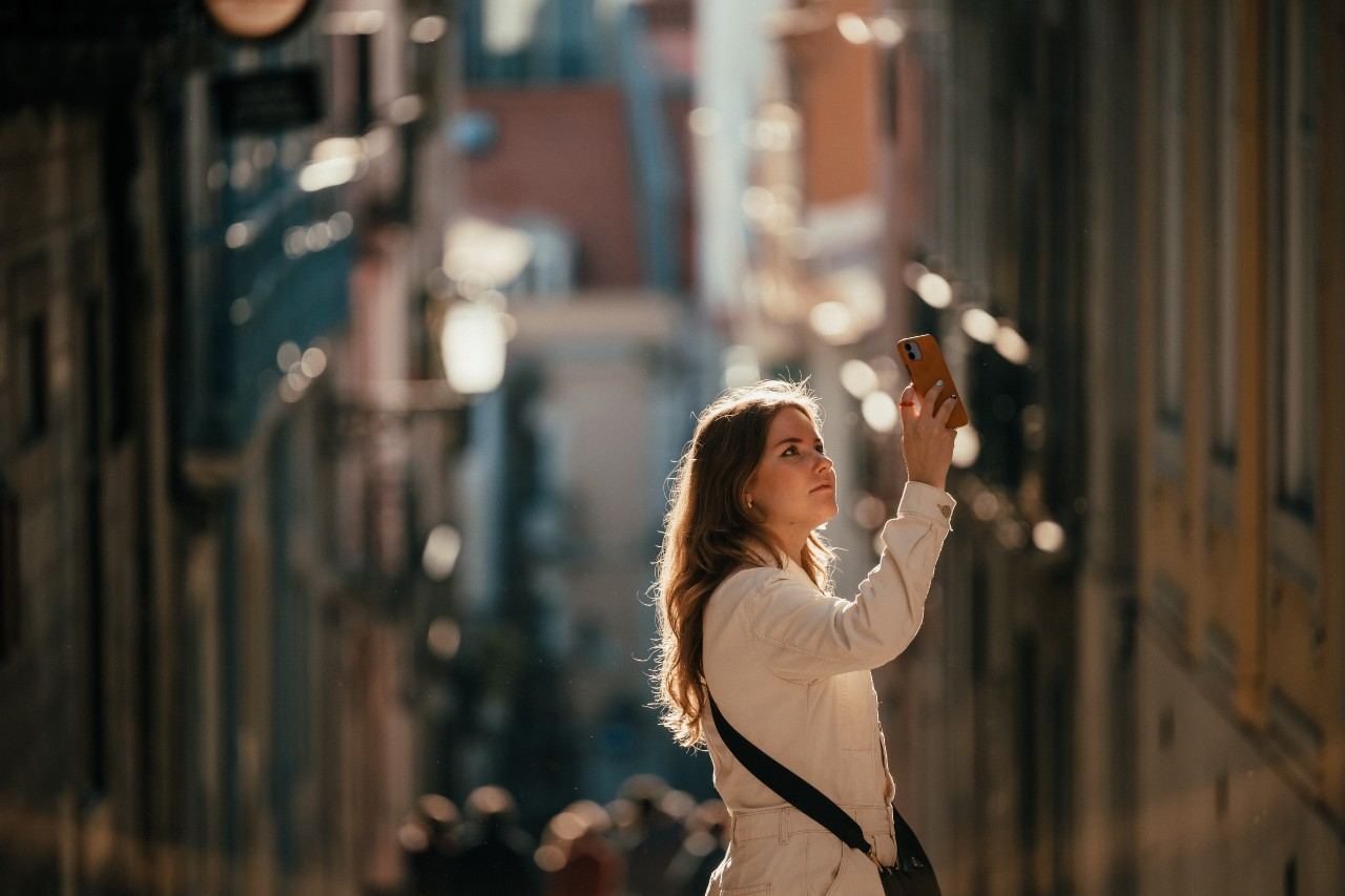 Junge Frau steht in einer sonnendurchfluteten Gasse und hält ihr Smartphone zum Fotografieren nach oben, wirkt konzentriert und neugierig. Junge Frau steht in einer sonnendurchfluteten Gasse und hält ihr Smartphone zum Fotografieren nach oben, wirkt konzentriert und neugierig.