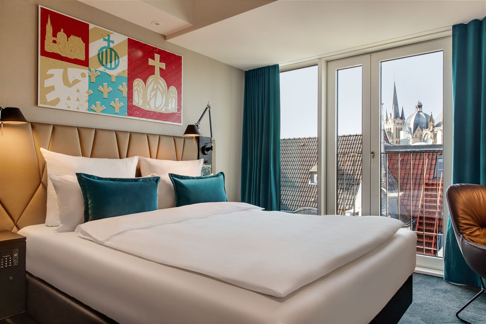 Helles Motel One Hotelzimmer mit großem, gemütlich bezogenem Doppelbett, moderner Wanddekoration und bodentiefen Fenstern mit Blick auf historische Kirchturmspitzen.