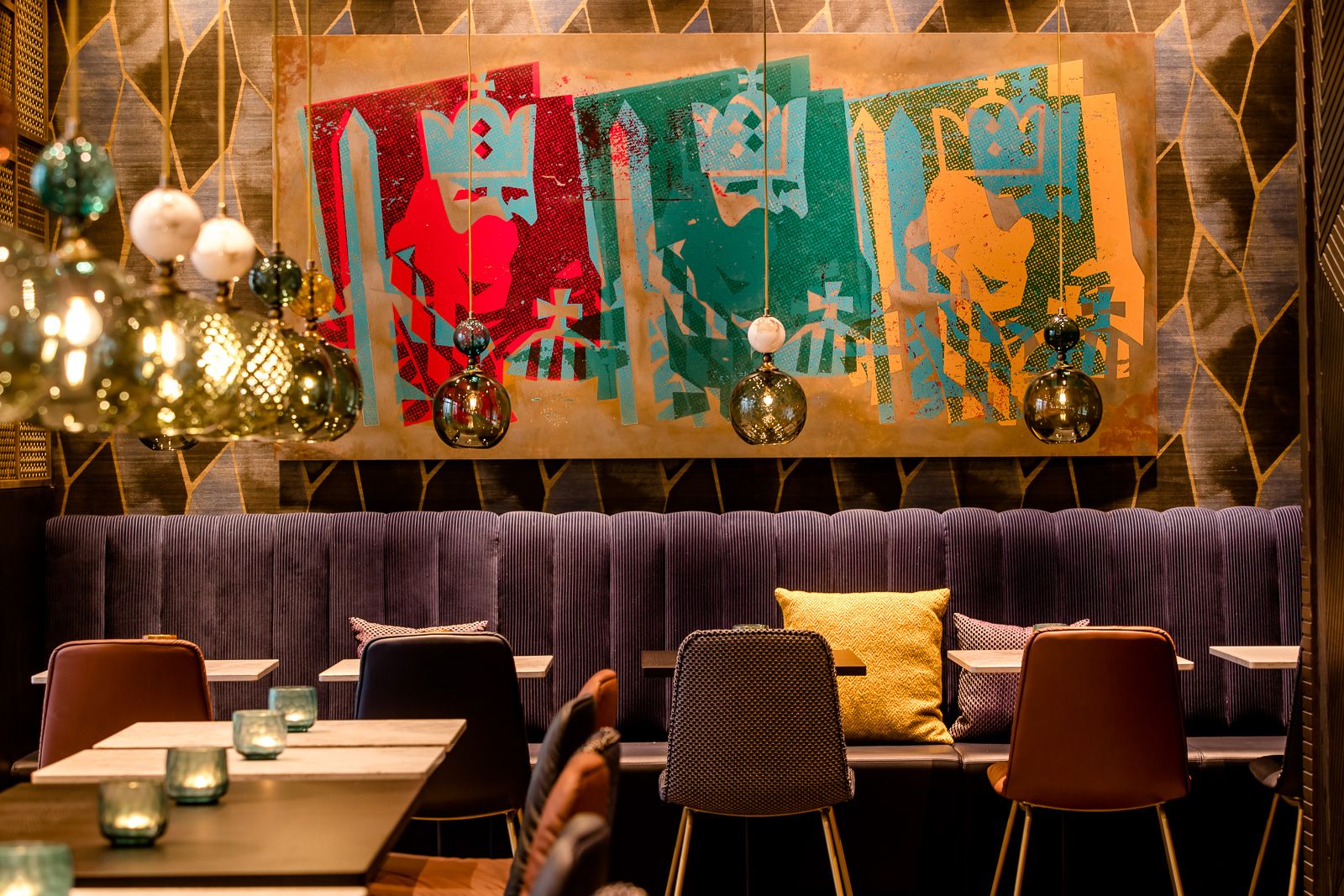 Stilvoll eingerichteter Motel One Hotel-Loungebereich mit samtigem Sitzsofa, farbenfroher moderner Wandkunst und eleganten Pendelleuchten, die warmes Licht über Tische und Stühle werfen. 