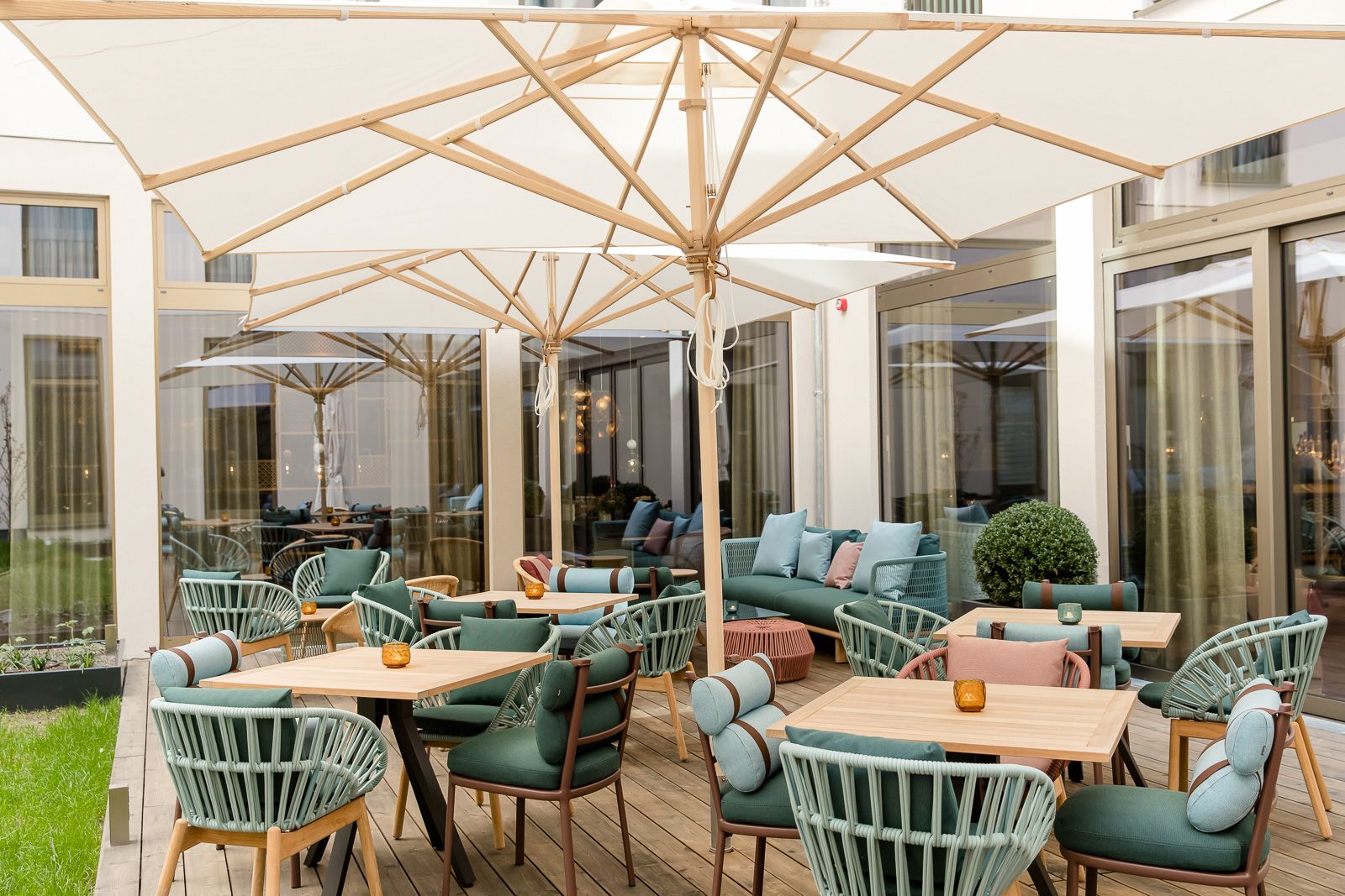 Helle Motel One Außenlounge mit Holzterrasse, modernen Stühlen und Sofas in Pastelltönen, großen Sonnenschirmen und Kerzenlicht.