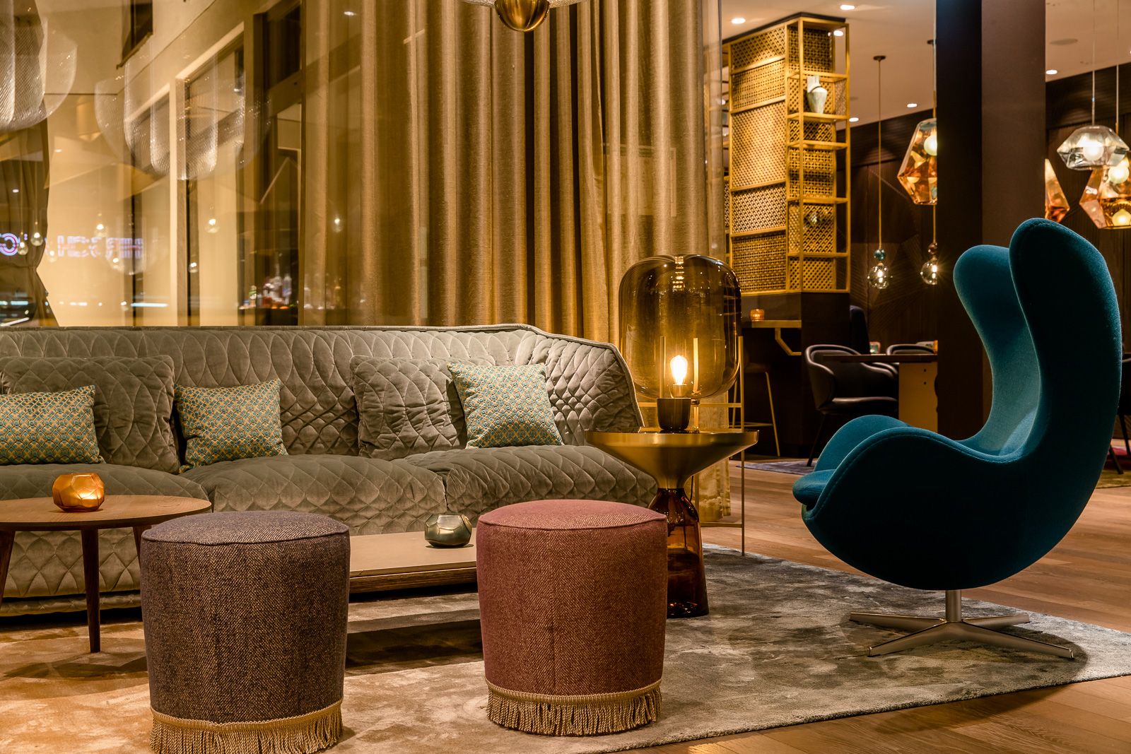 Gemütliche Motel One Lounge mit stilvollem, grauem Sofa, bunten Sitzhockern und einem modernen, blauen Design-Sessel. 