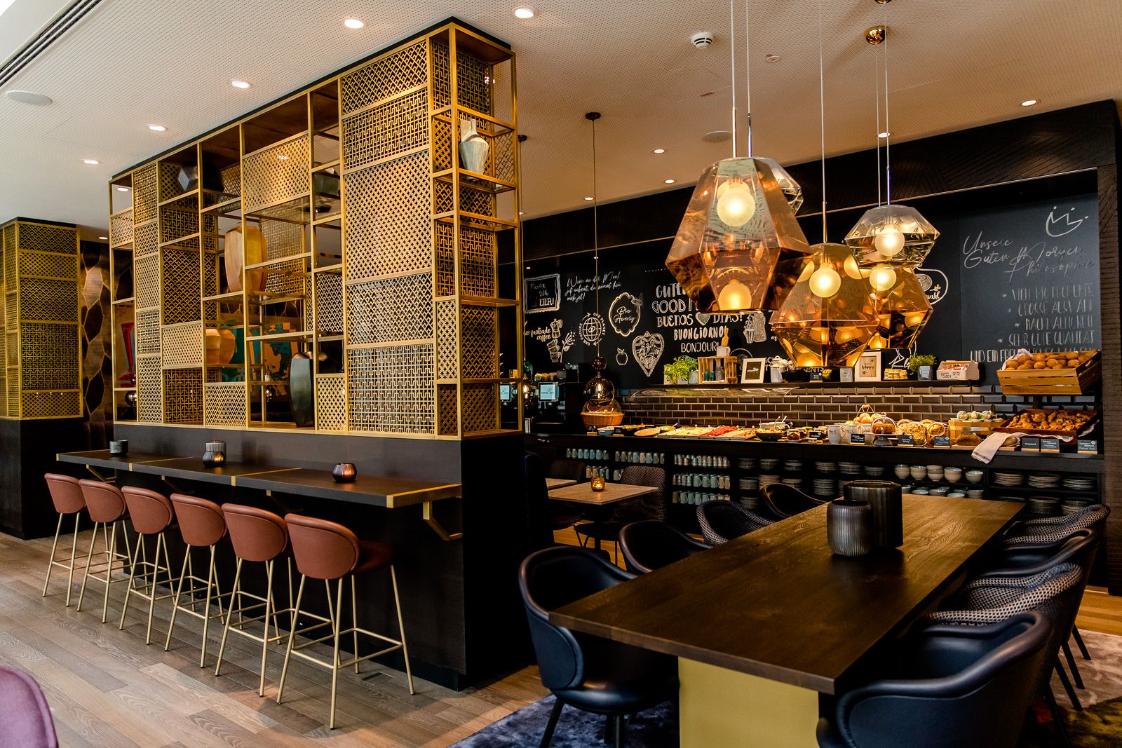 Modern eingerichteter Motel One Frühstücksraum mit langen Tischen, bequemen Stühlen und stilvollen Pendelleuchten, Buffetbereich mit frischen Backwaren und Getränken