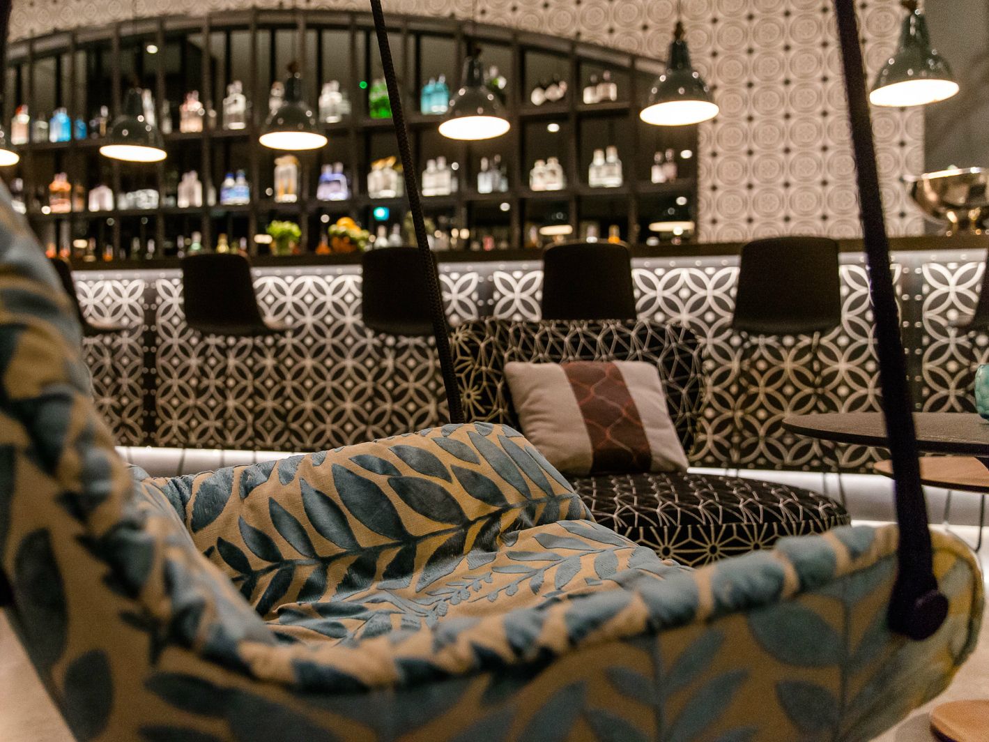 Lounge-Bereich mit bequemen, gemusterten Sesseln und Kissen, stimmungsvollem Licht und einer gut bestückten Bar im Hintergrund. 