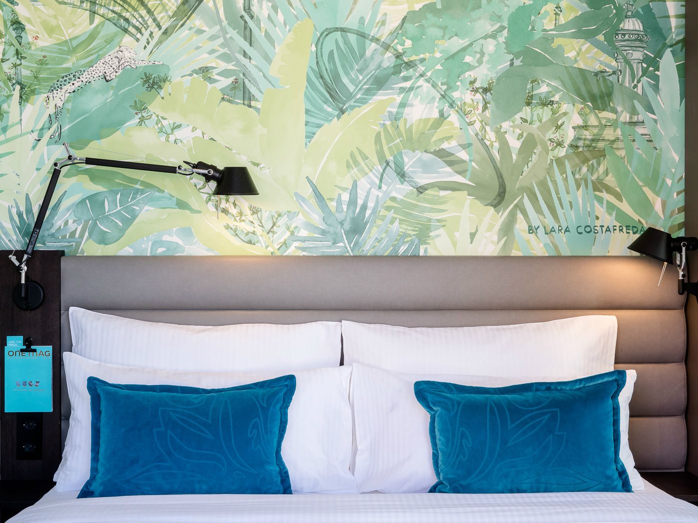 Modernes Hotelbett mit weißer Bettwäsche und zwei türkisen Kissen vor einer großen Wand mit exotischem, grünem Pflanzenmuster, kombiniert mit Leselampen für gemütliche Atmosphäre.