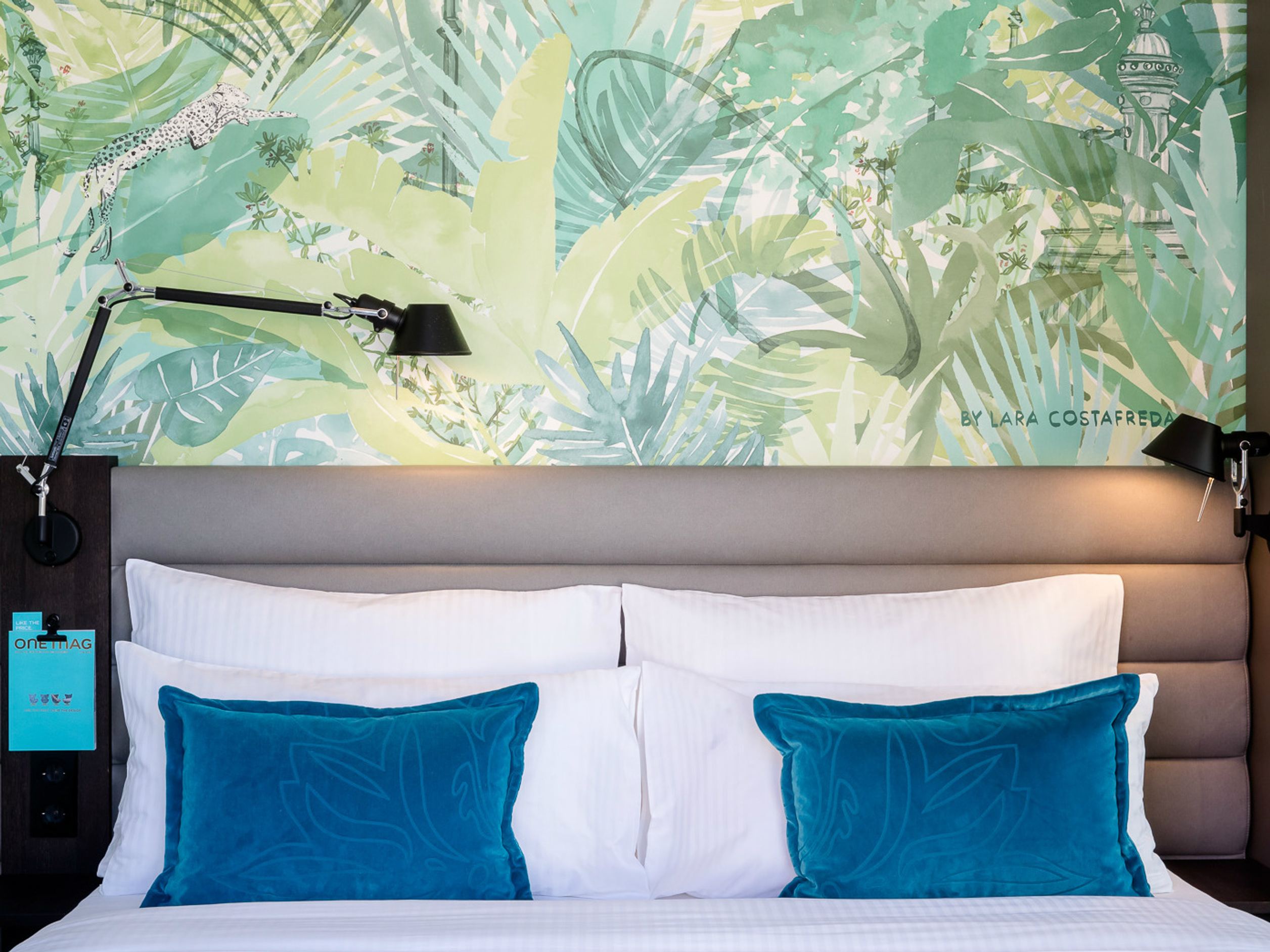 Motel One Doppelbett mit weißer Bettwäsche und zwei türkisen Kissen vor einer großen Wand mit exotischem, grünem Pflanzenmuster, kombiniert mit Leselampen.