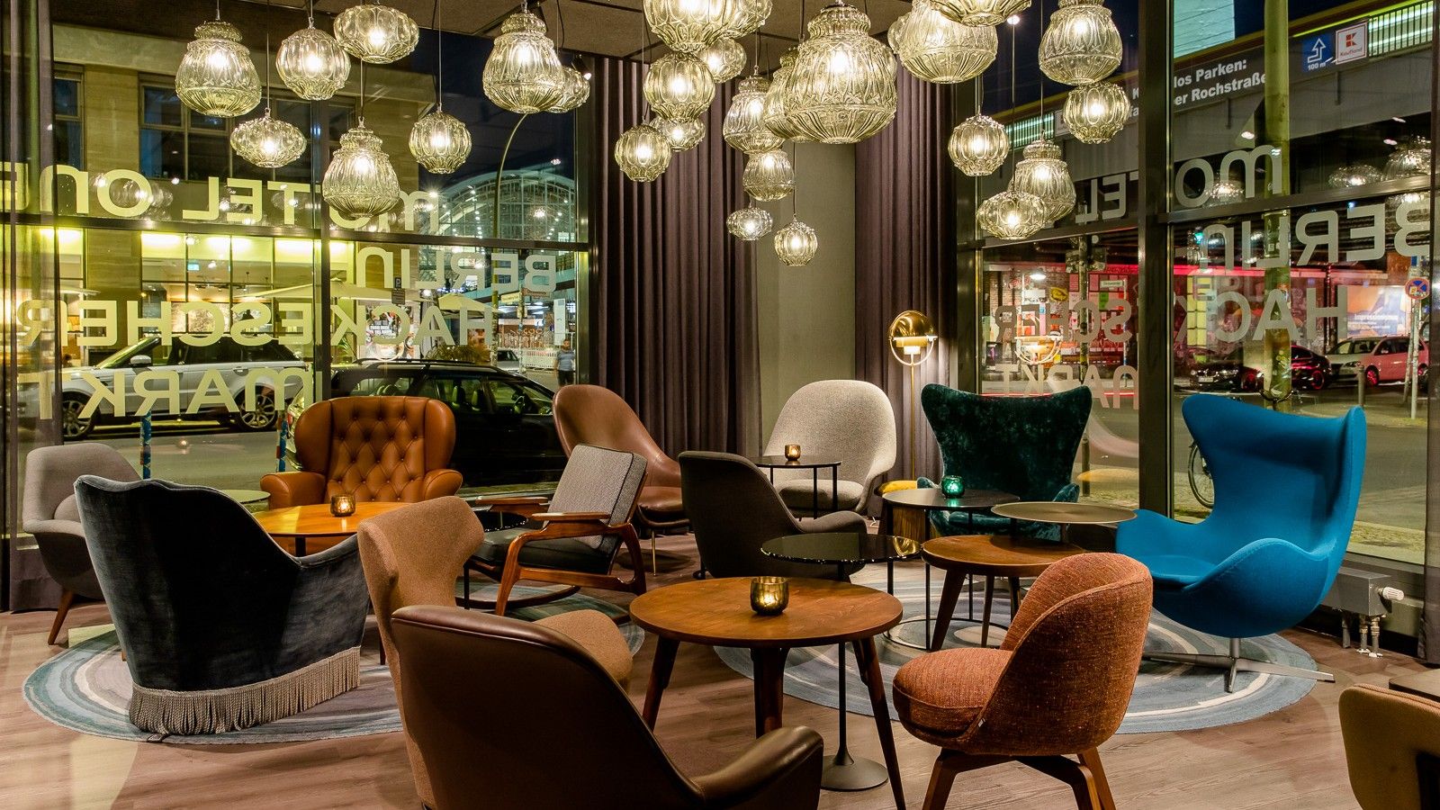 Gemütliche Hotel-Lounge mit modernen, farbenfrohen Sesseln und warmem Licht durch zahlreiche hängende Lampen, großer Fensterfront mit Blick auf eine beleuchtete Straße.