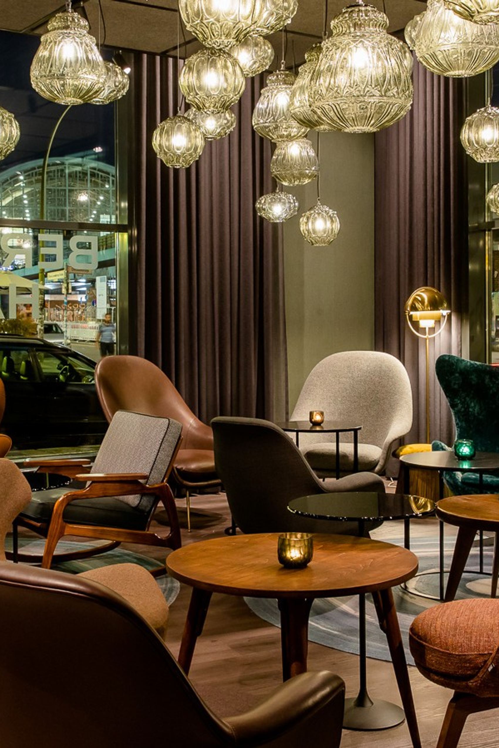 Gemütliche Hotel-Lounge mit modernen, farbenfrohen Sesseln und warmem Licht durch zahlreiche hängende Lampen, großer Fensterfront mit Blick auf eine beleuchtete Straße.