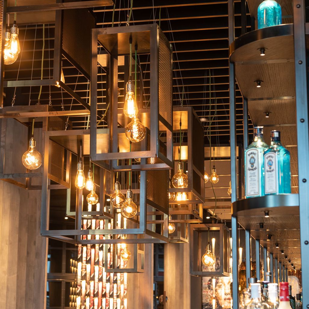 Moderne Bar mit warm beleuchteten Vintage-Glühbirnen in geometrischen Metallrahmen, die eine einladende und stilvolle Atmosphäre schaffen. Regale mit dekorativen Flaschen und Holzvertäfelung ergänzen das urbane Design.