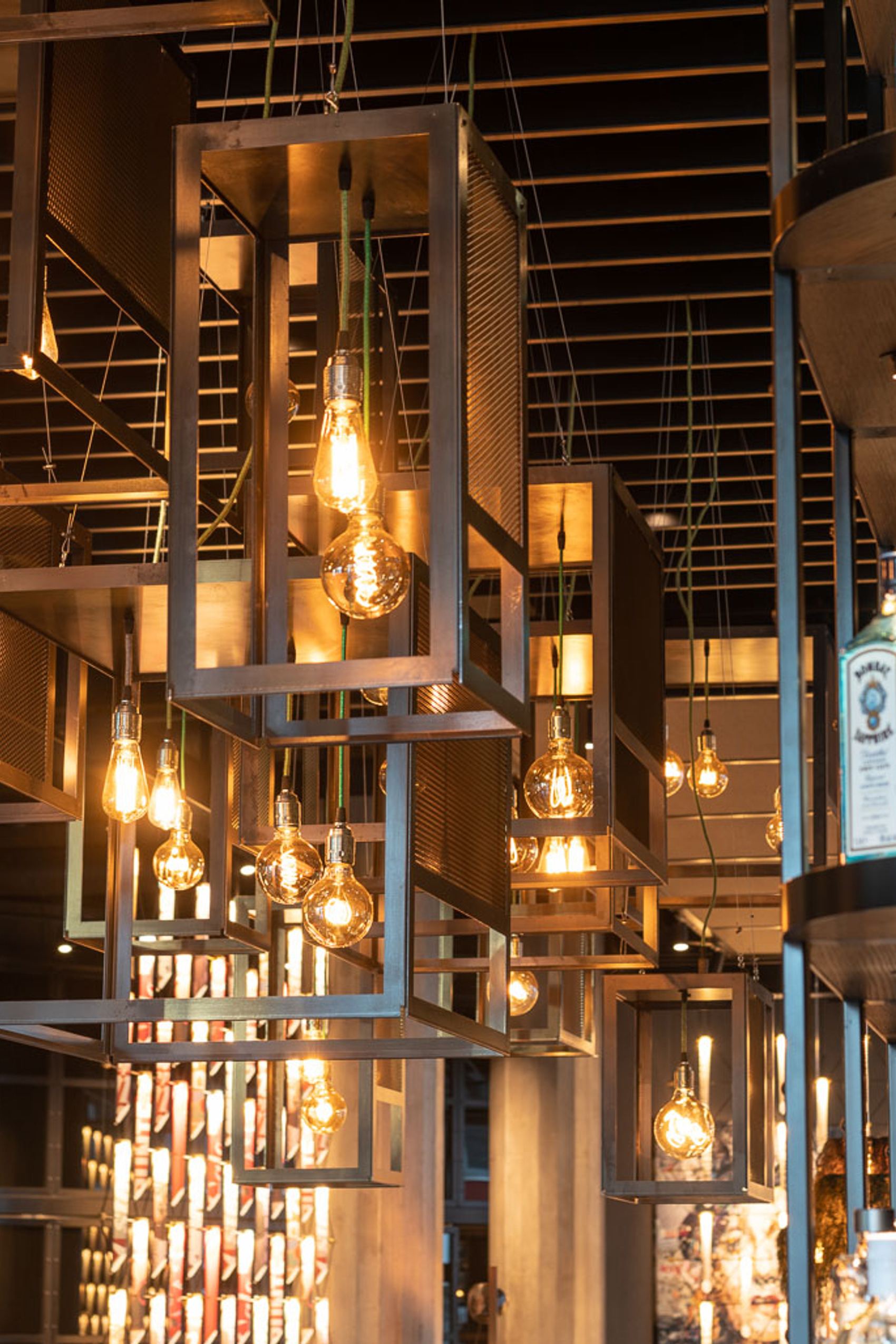 Moderne Bar mit warm beleuchteten Vintage-Glühbirnen in geometrischen Metallrahmen, die eine einladende und stilvolle Atmosphäre schaffen. Regale mit dekorativen Flaschen und Holzvertäfelung ergänzen das urbane Design.