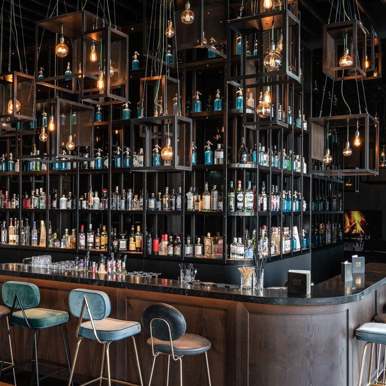 Moderne Hotelbar mit dunklem Holz, hohen Regalen voller Spirituosen, stilvollen Hängelampen und verschiedenen Barhockern. 