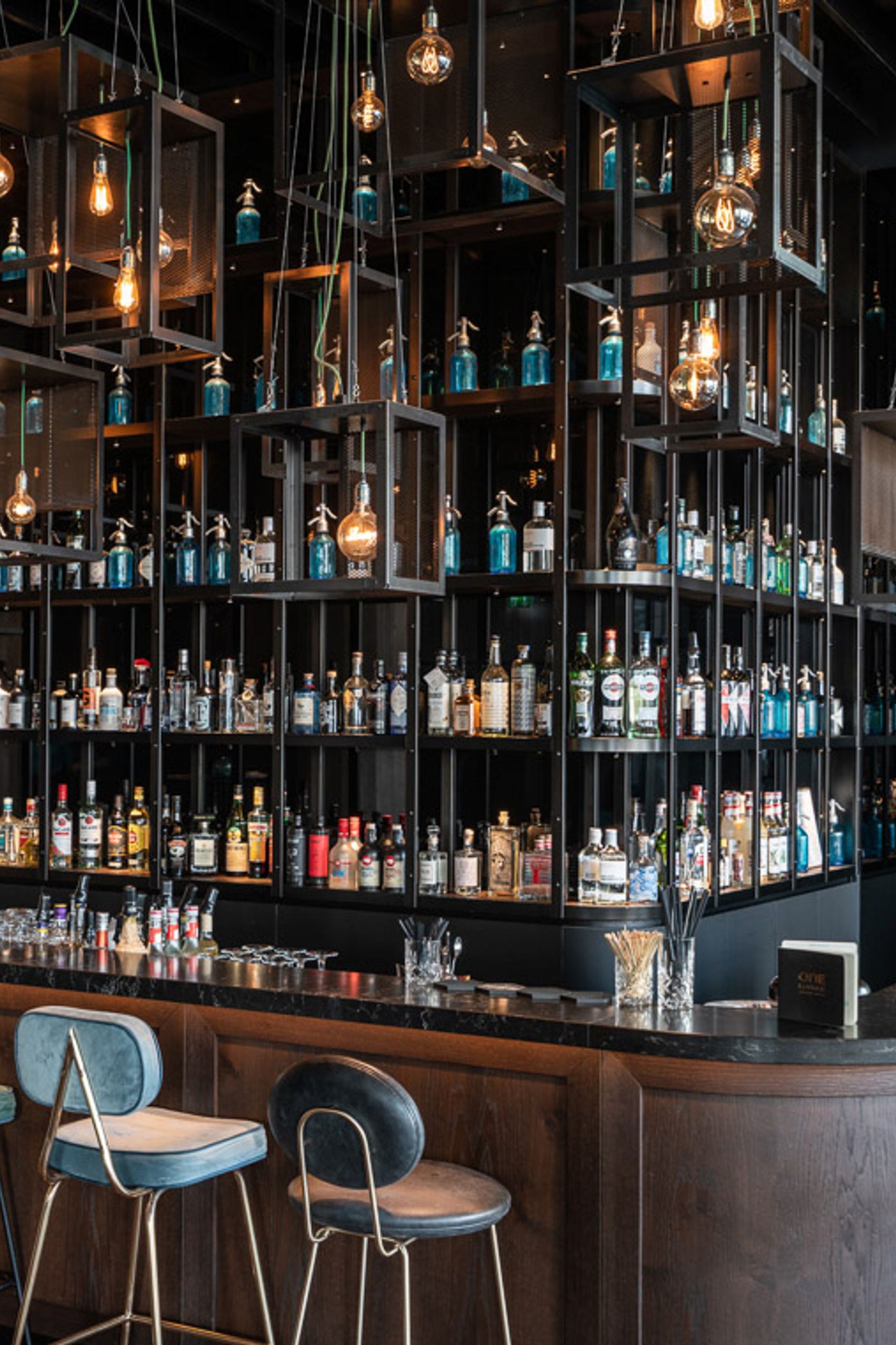 Motel One Hotelbar mit dunklem Holz, hohen Regalen voller Spirituosen, stilvollen Hängelampen und verschiedenen Barhockern.