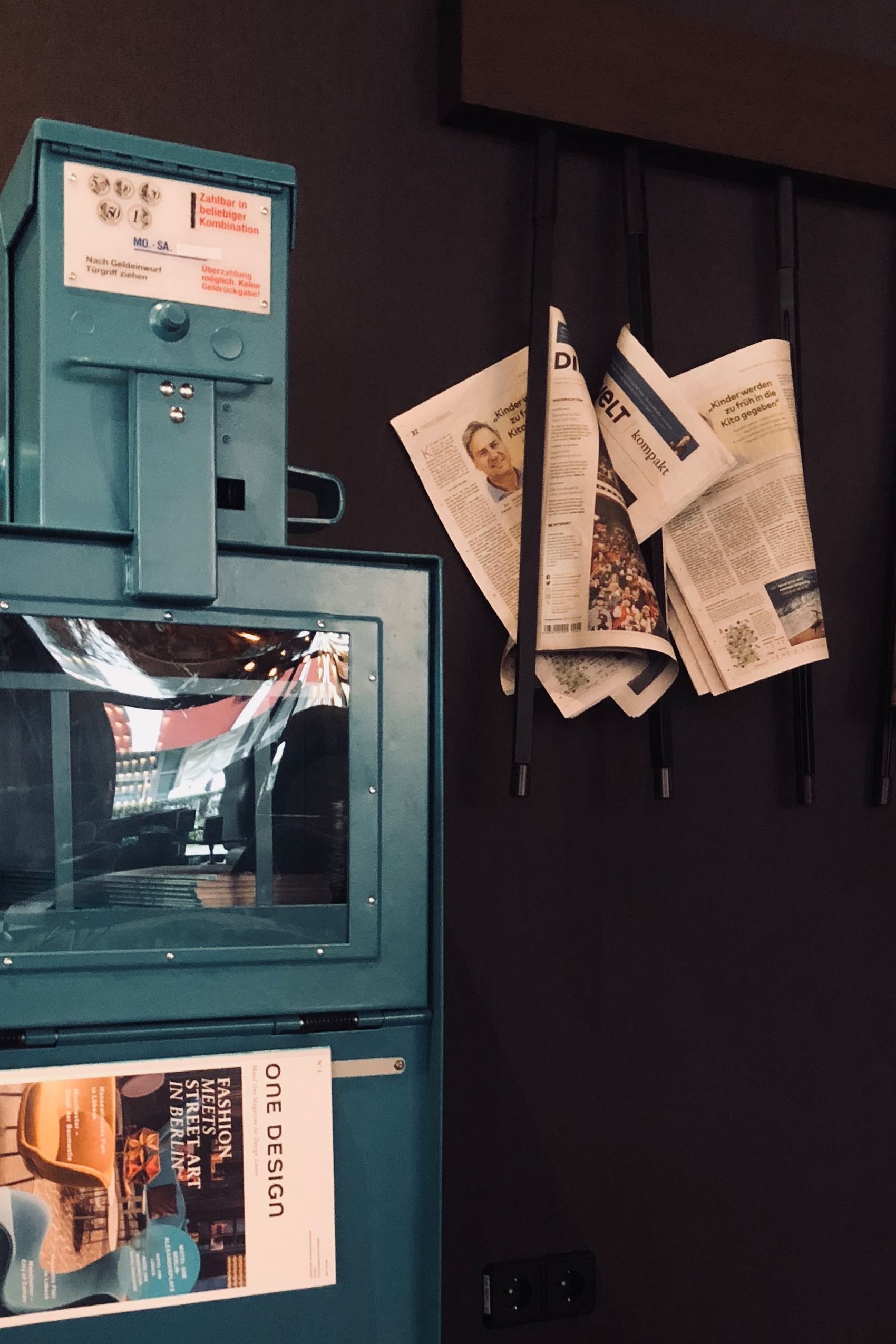 Blauer Zeitungshalter mit einem Magazin vorne, daneben an der Wand vier Zeitungen, die in einem schwarzen Halter hängen, vor dunkelgrauem Hintergrund.
