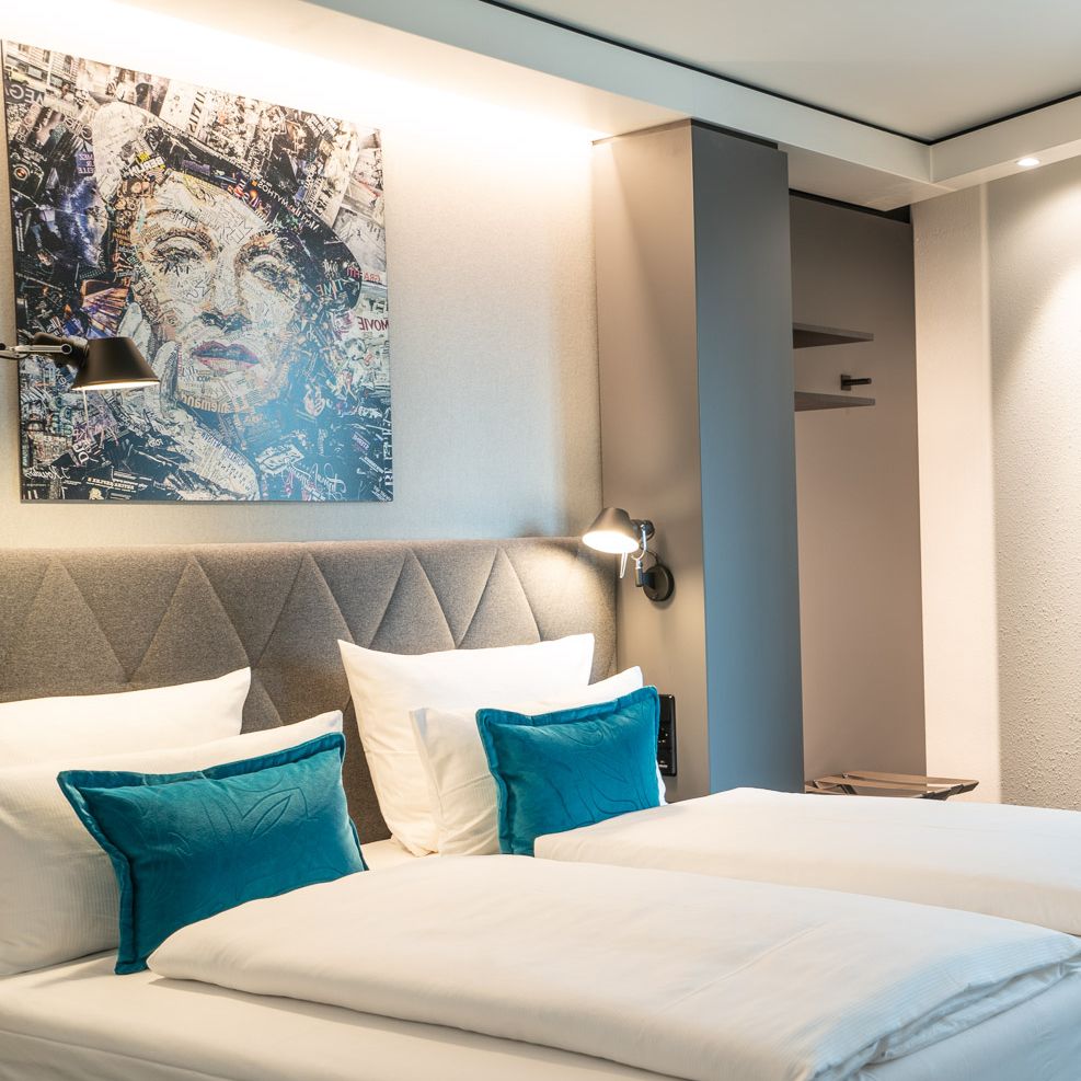 Modernes Hotelzimmer mit zwei Einzelbetten, weißer Bettwäsche und türkisfarbenen Kissen. Dezente Beleuchtung und modernes Kunstbild über dem gepolsterten Kopfteil.