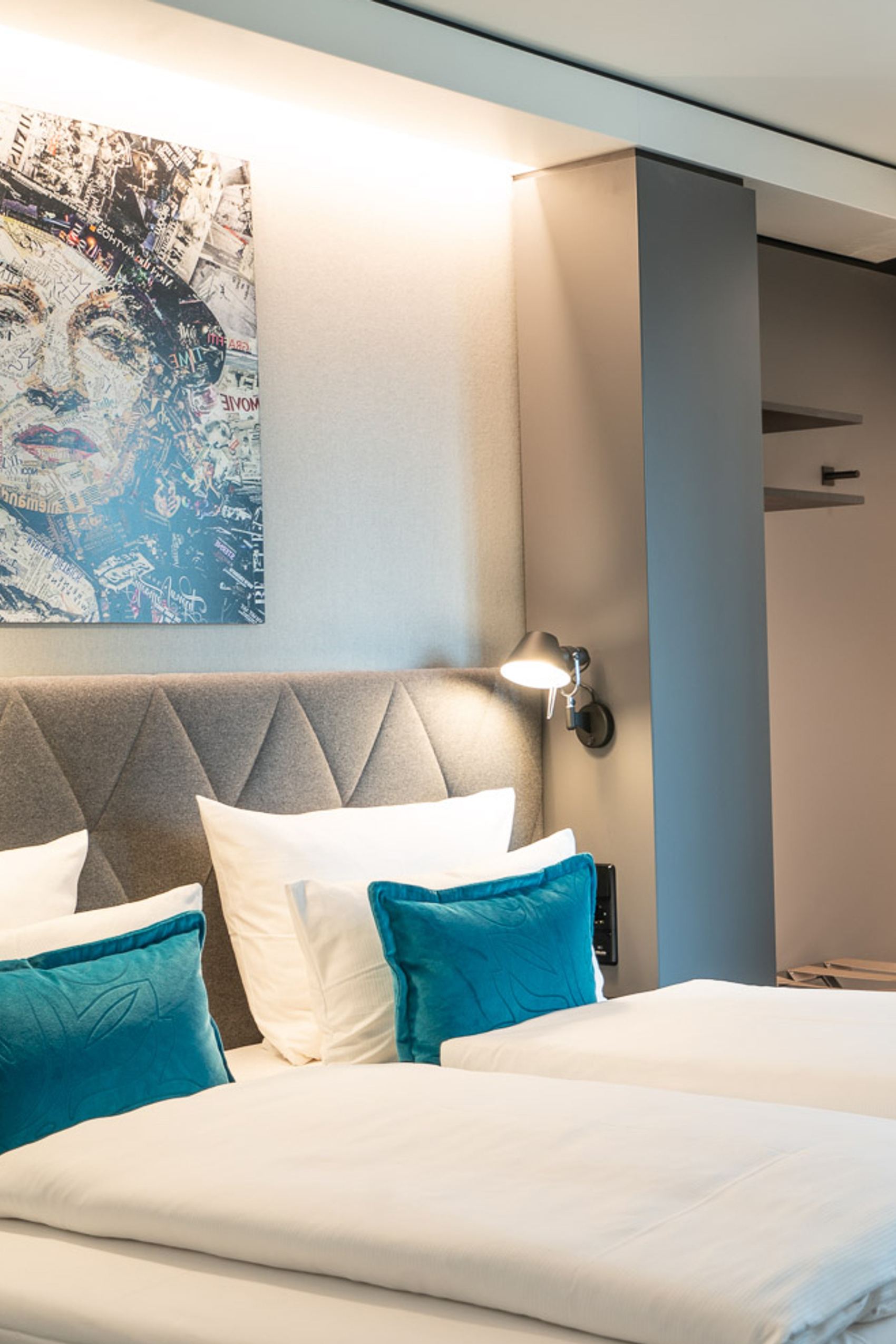 Motel One Hotelzimmer mit zwei Einzelbetten, weißer Bettwäsche und türkisfarbenen Kissen. Dezente Beleuchtung und modernes Kunstbild über dem gepolsterten Kopfteil.