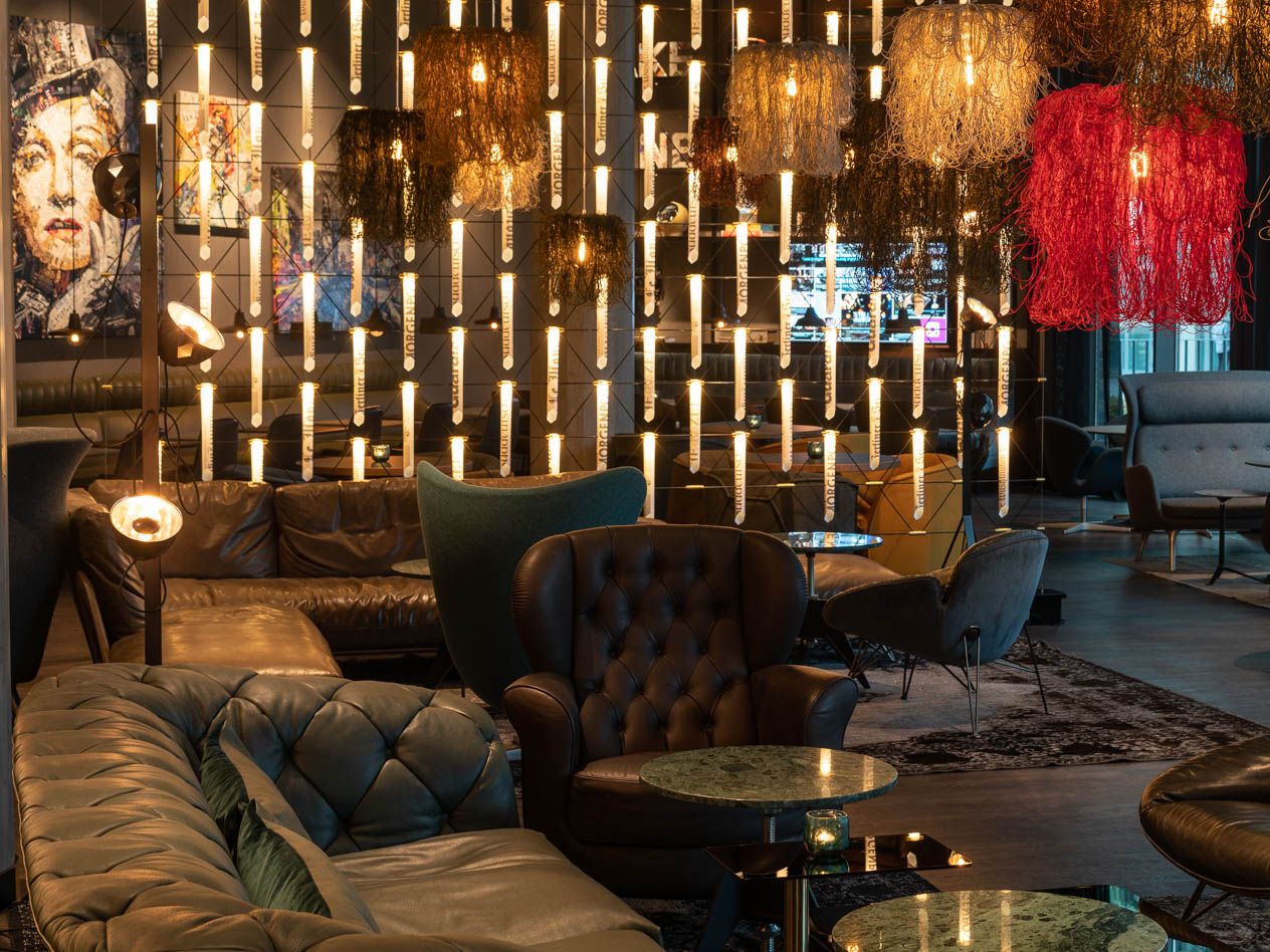 Gemütliche Lounge mit Ledersesseln und Sofas, umgeben von warmem, kreativem Lichtdesign mit zahlreichen hängenden Leuchten. 