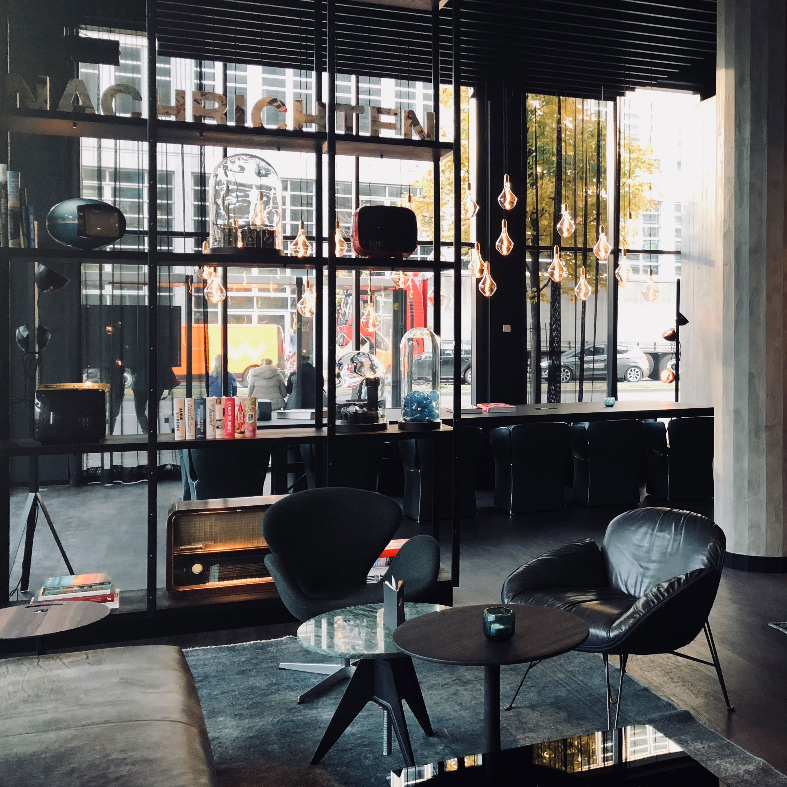 Modern gestalteter Loungebereich mit Sitzgelegenheiten, stilvollen Glühbirnen und großem Fenster, das Tageslicht hereinlässt. Gemütliche Atmosphäre mit retro-inspirierten Möbeln und dekorativen Elementen, ideal zum Entspannen oder Arbeiten bei natürlichem Licht.