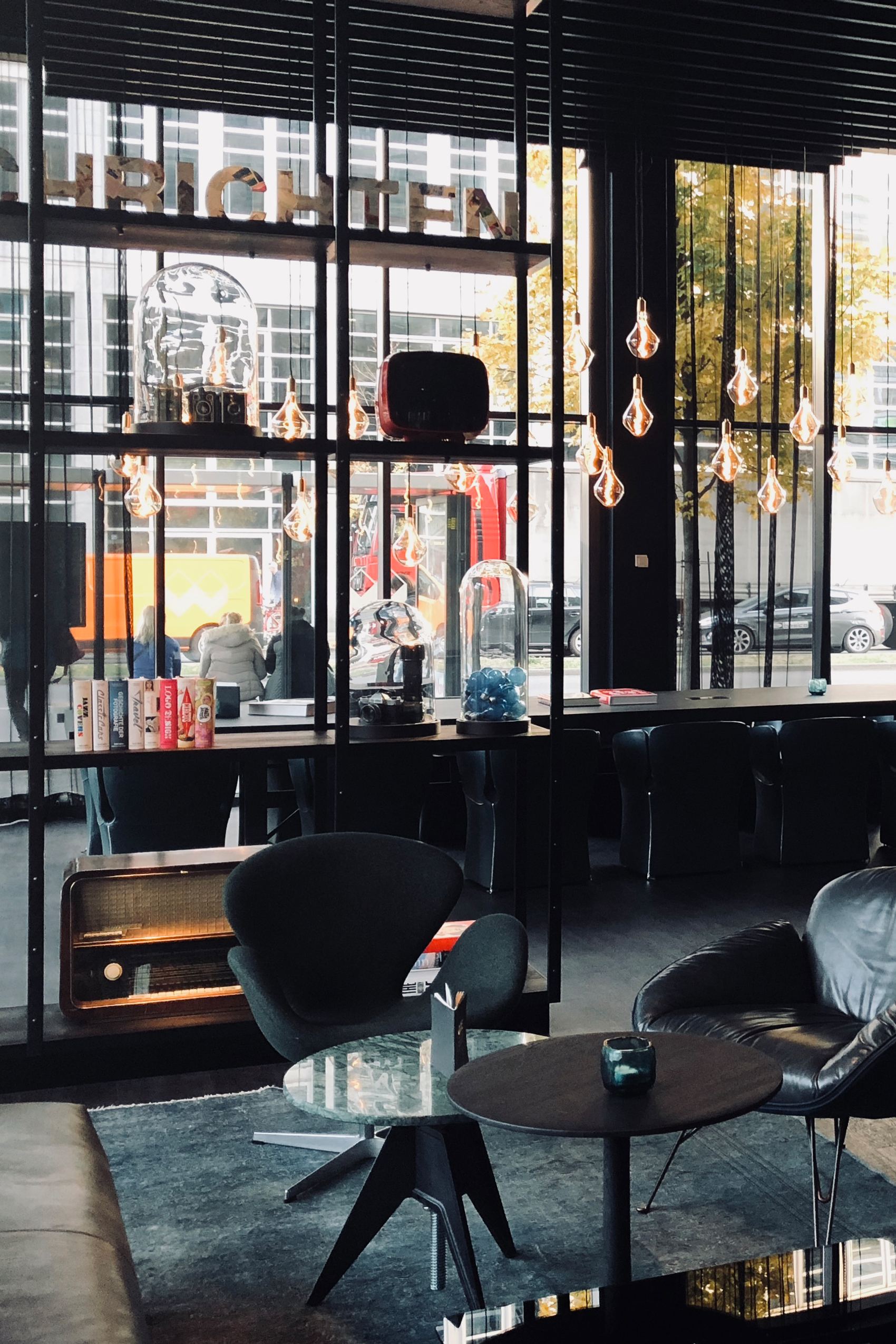 Modern gestalteter Loungebereich mit Sitzgelegenheiten, stilvollen Glühbirnen und großem Fenster, das Tageslicht hereinlässt. Gemütliche Atmosphäre mit retro-inspirierten Möbeln und dekorativen Elementen, ideal zum Entspannen oder Arbeiten bei natürlichem Licht.