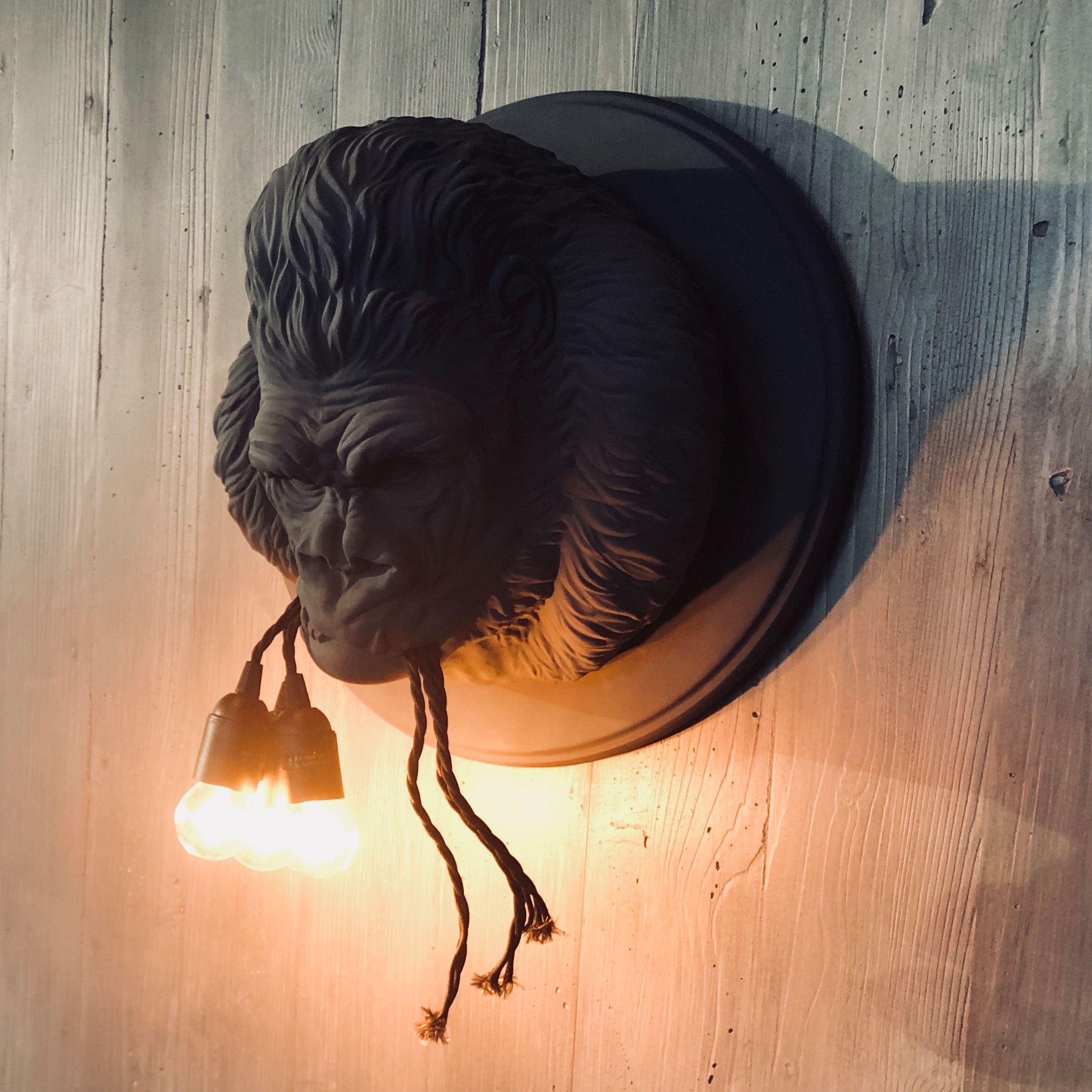 Wandlampe in Form eines detailierten Gorilla-Kopfes, aus dessen Mund zwei leuchtende Glühbirnen hängen. Rustikaler Holzuntergrund betont die warme, atmosphärische Beleuchtung, ideal für besondere Raumakzente.
