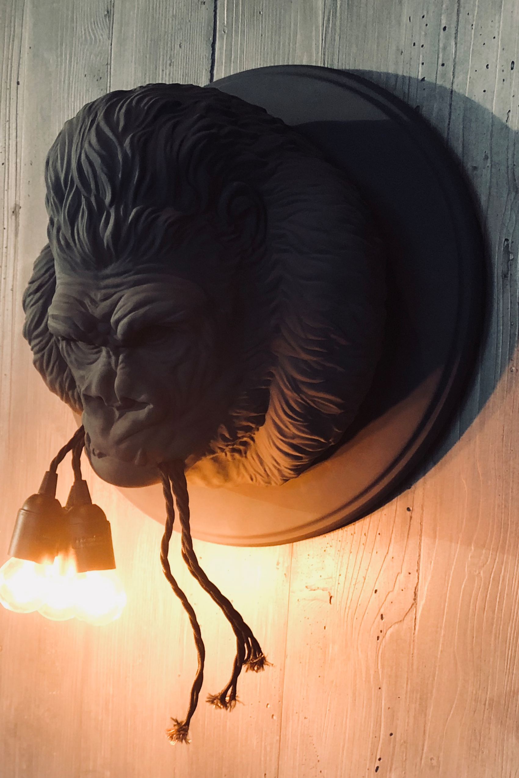 Wandlampe in Form eines detailierten Gorilla-Kopfes, aus dessen Mund zwei leuchtende Glühbirnen hängen. Rustikaler Holzuntergrund betont die warme, atmosphärische Beleuchtung, ideal für besondere Raumakzente.