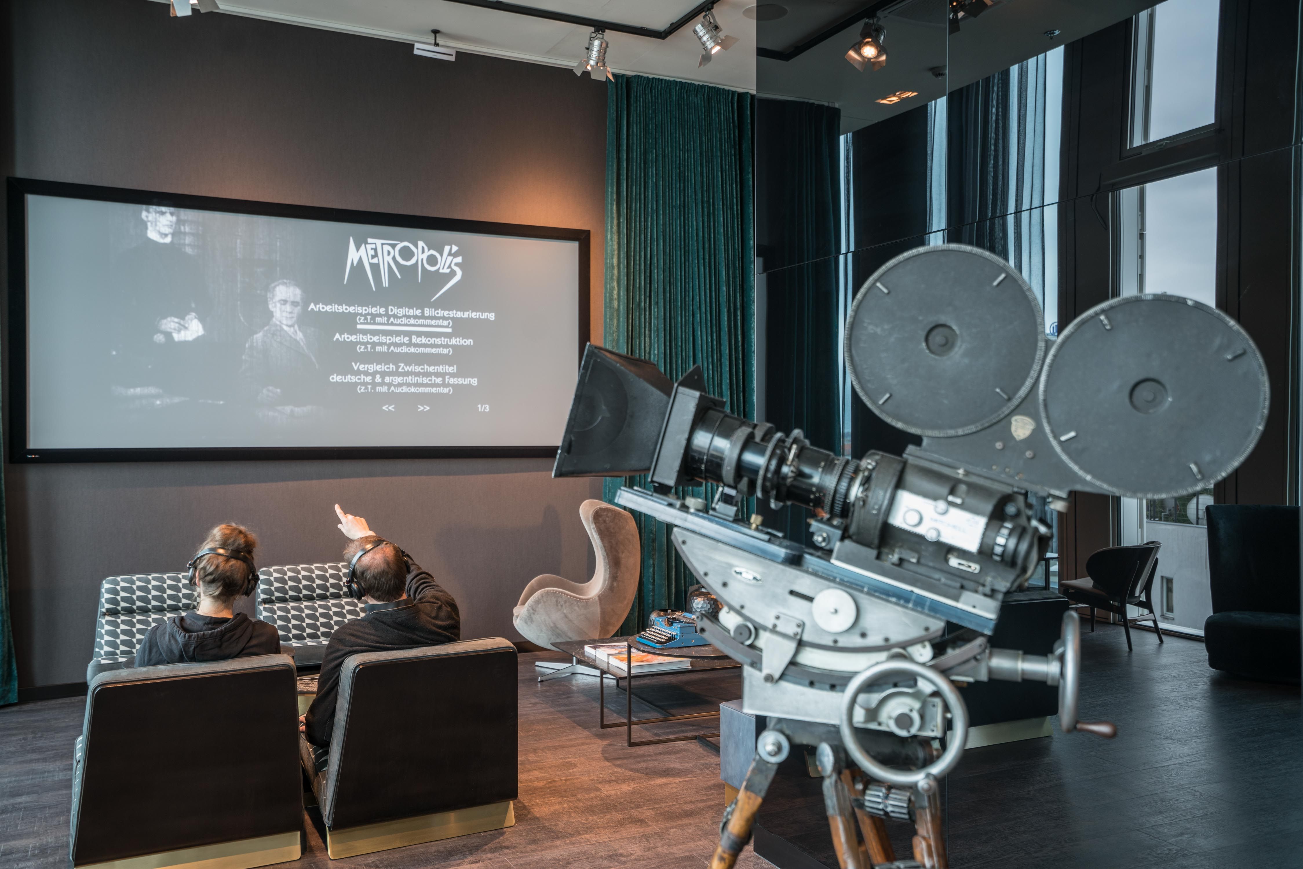 Zwei Personen sehen in einem modernen Design Kinoraum im Motel One einen Film auf großer Leinwand. Im Vordergrund steht eine vintage Filmkamera, die Atmosphäre verbindet Filmkunst mit zeitgemäßem Komfort.