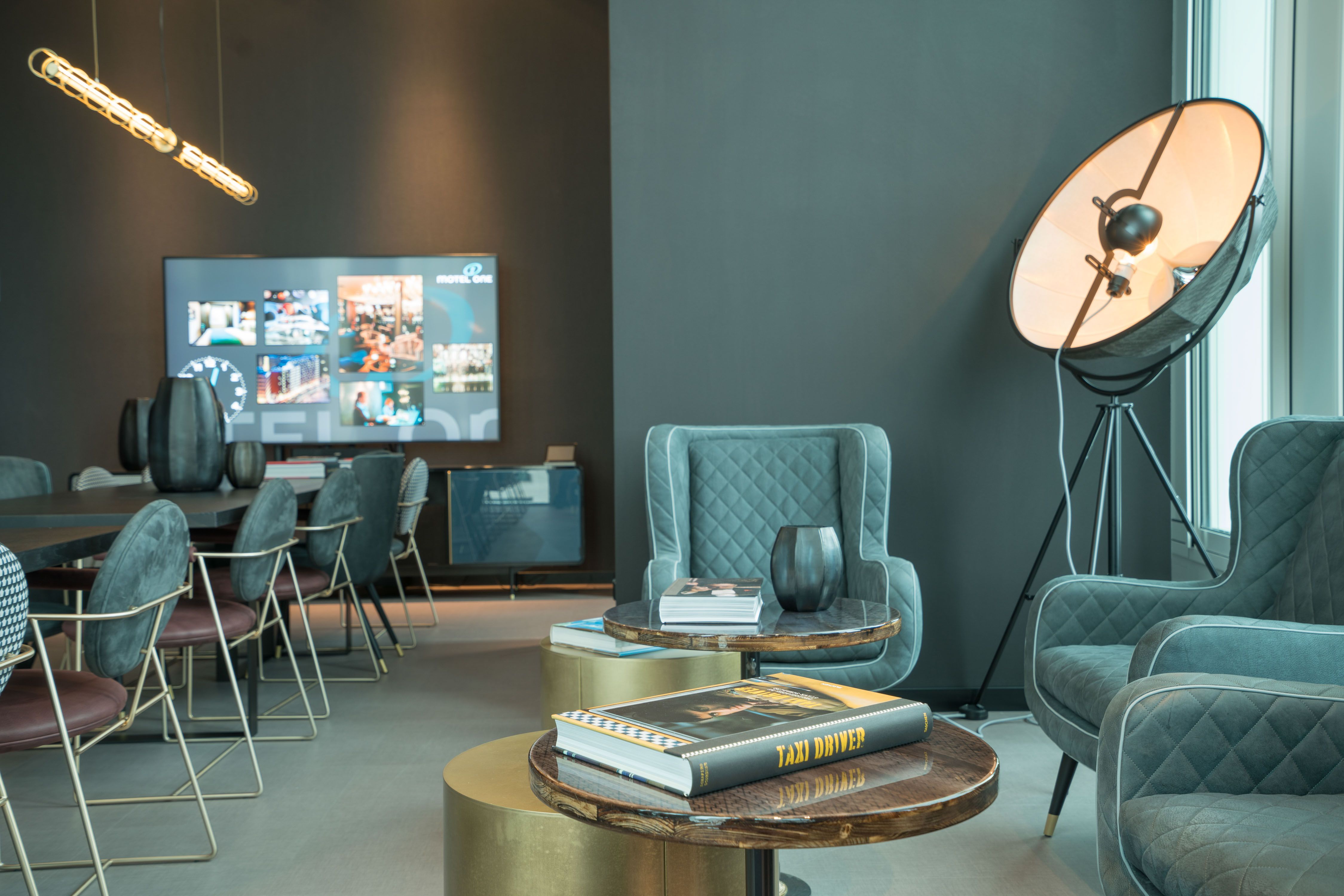 Moderne Lounge mit bequemen Sesseln, kleinen Tischen und stilvoller Beleuchtung, ideal zum Entspannen oder für informelle Gespräche. Bücher und Dekor auf den Tischen betonen gemütliche Atmosphäre. Im Hintergrund ein großer Bildschirm und Esstische für Arbeit oder Meetings.
