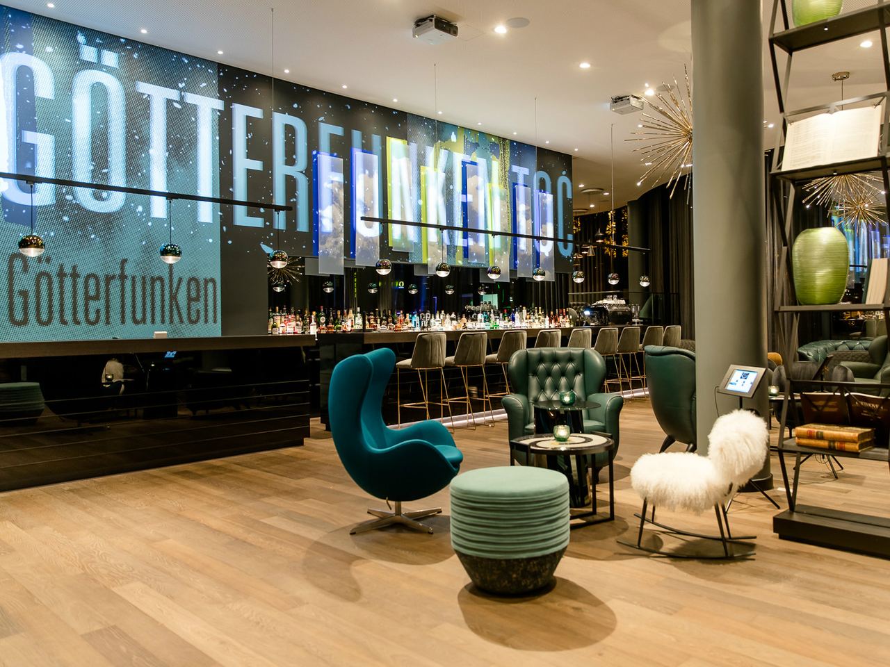 Motel One Hotelbar mit stilvoller Beleuchtung, gemütlichen Sitzgelegenheiten und einer langen Theke mit Barhockern. Großes, beleuchtetes Schriftzug-Display an der Wand.
