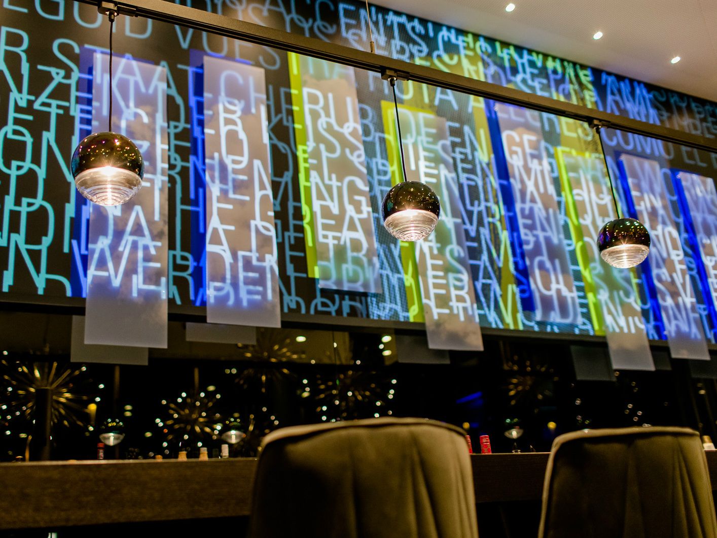 Bunte, beleuchtete Wand mit überlagerten Buchstaben und modernen Hängelampen in einem stilvollen Innenraum, der eine warme und einladende Atmosphäre schafft.