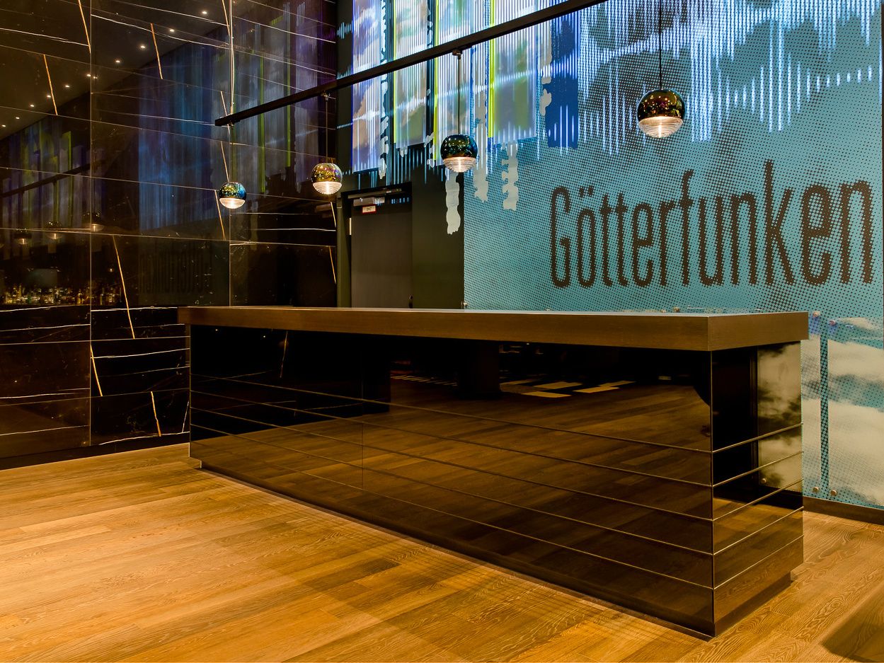 Moderne Motel One Hotelrezeption mit glänzend schwarzem Tresen, warmem Holzboden und dekorativer Beleuchtung vor einer Wand mit beleuchtetem Schriftzug „Götterfunken“. 