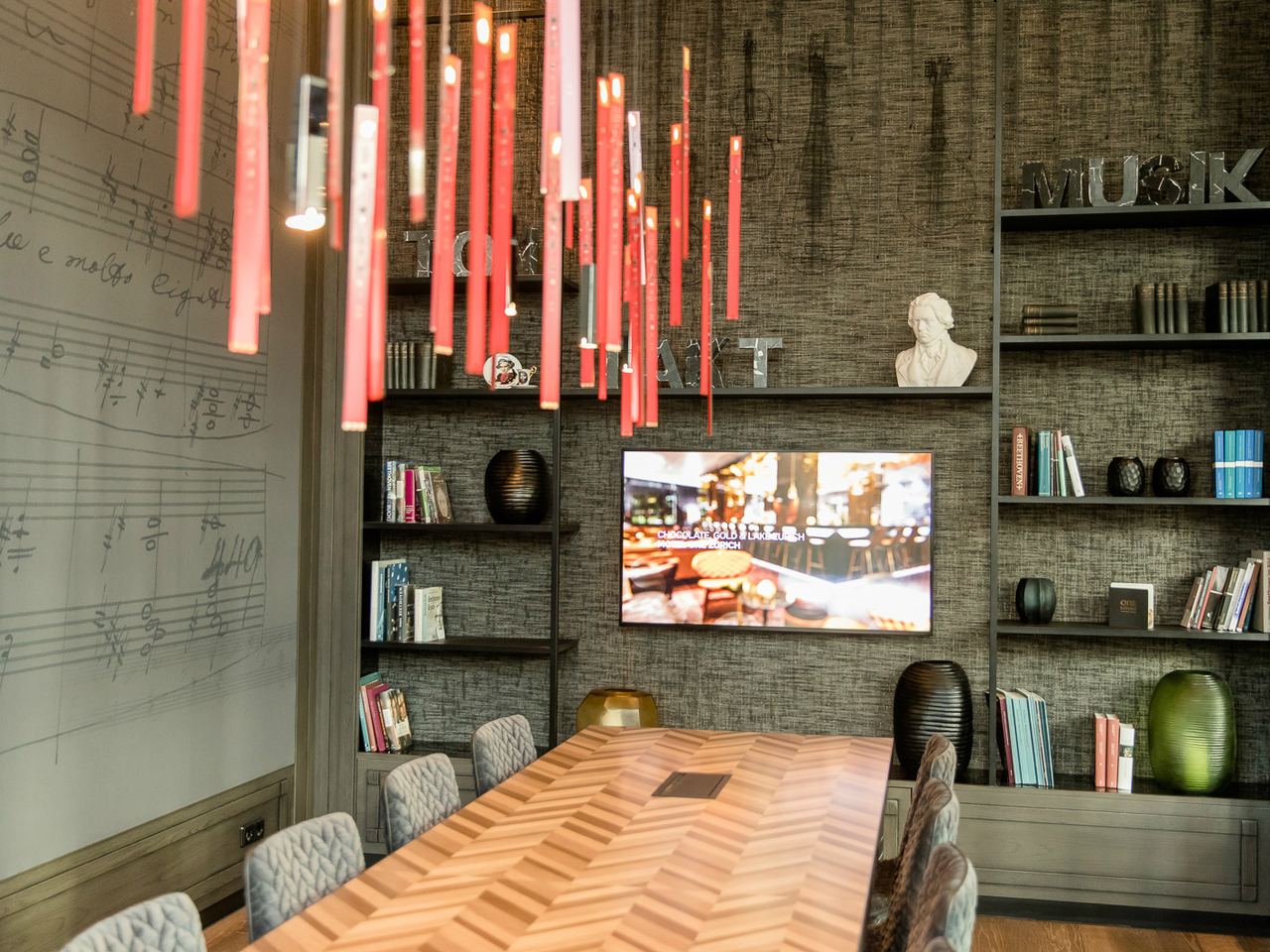Moderner Besprechungsraum mit langem Holztisch, gepolsterten Stühlen und dekorativer Wandbeleuchtung. Bücherregale und Musiknoten an der Wand schaffen eine kreative und inspirierende Atmosphäre. Bildschirm für Präsentationen ist integriert.