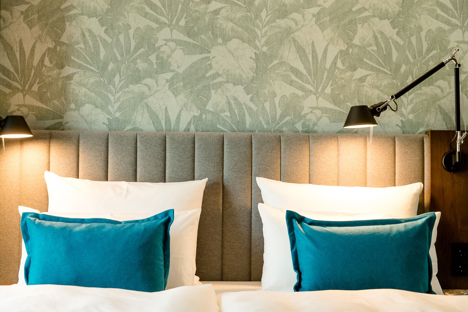Doppelbett mit grauem, gepolstertem Kopfteil, zwei weißen Kissen und je einem türkisfarbenen Kissen, umgeben von warmem Licht und einer grünen, pflanzenartigen Tapete im Hintergrund.