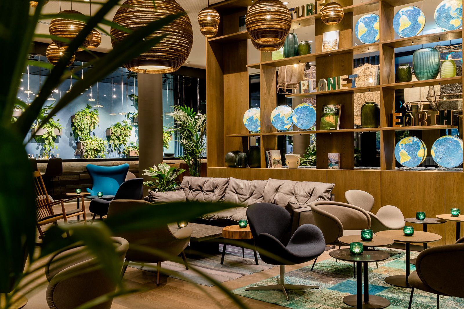 Gemütliche Lounge mit bequemen Sesseln, Sofas und grünen Pflanzen. Warme Holzelemente und dekorative Globus-Modelle schaffen eine einladende, entspannte Atmosphäre ideal zum Verweilen nach einem langen Reisetag.