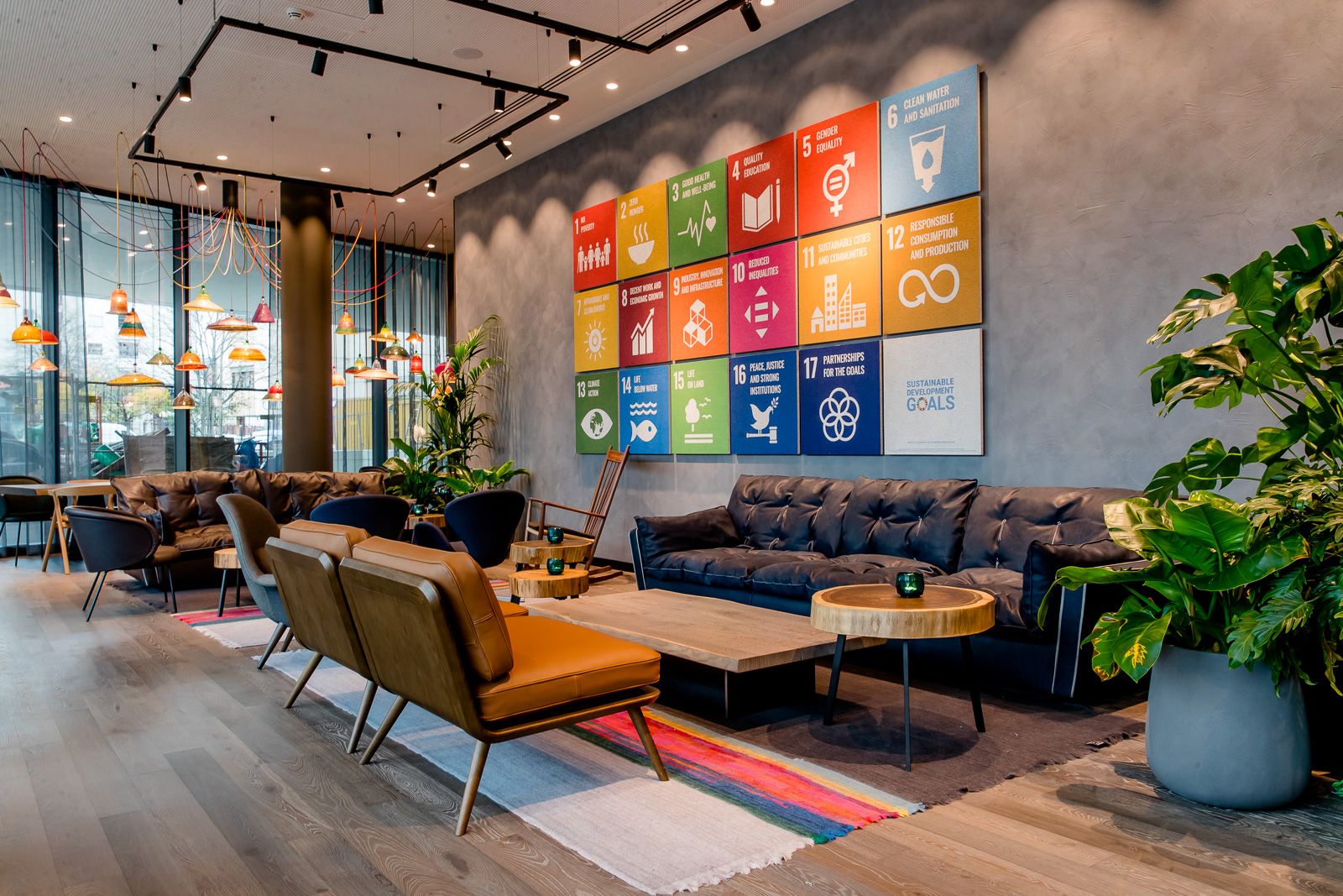 Gemütlicher Lounge-Bereich mit modernen Sitzmöbeln und Pflanzen, an der Wand bunte Tafeln mit den 17 Zielen für nachhaltige Entwicklung der Vereinten Nationen. Großflächige Fenster lassen viel Tageslicht herein.