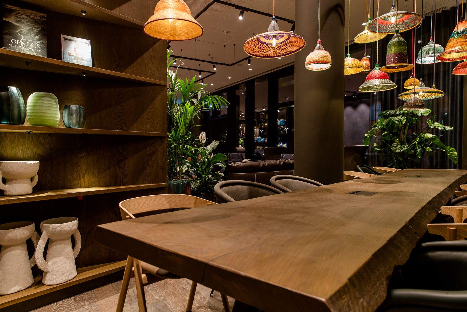 Moderner Essbereich mit großem Holztisch, stilvollen Stühlen und hängenden Lampenschirmen in verschiedenen Farben, umgeben von grünen Pflanzen 