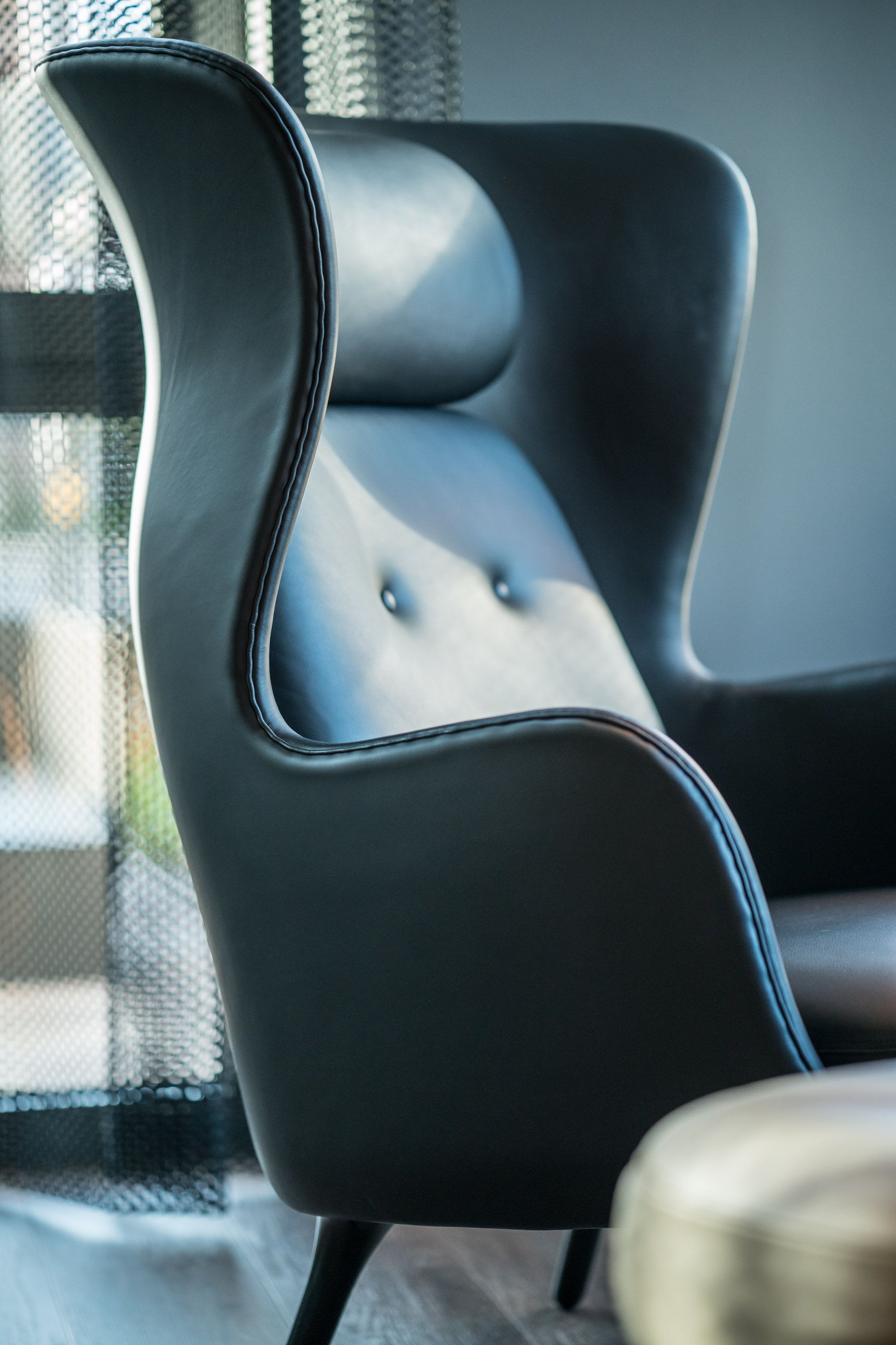 Eleganter, hoher Ledersessel mit geschwungenem Design, komfortabler Polsterung und dezenter Knopfverzierung in einem modernen, ruhigen Raum.