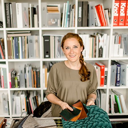 Frau in hellem Raum hält verschiedene Stoffproben, umgeben von Regalen mit Ordnern und Büchern, wirkt freundlich und engagiert.