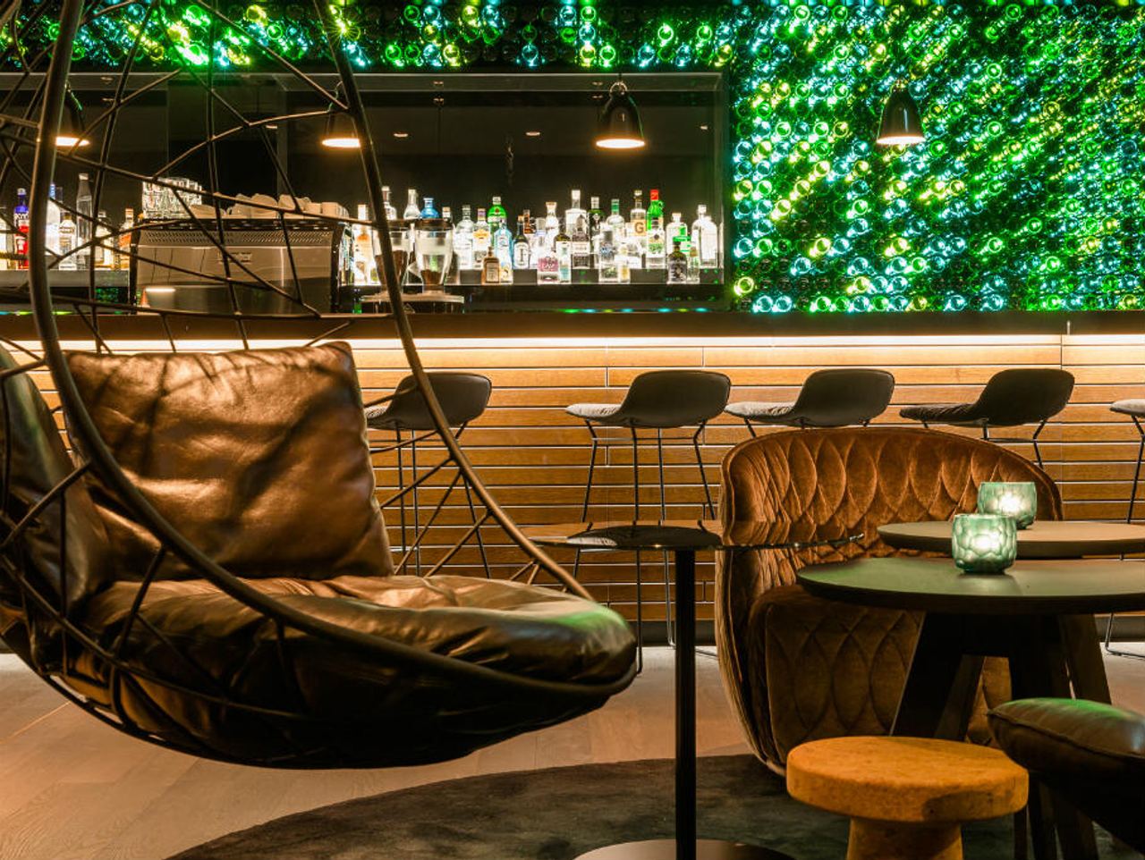 Gemütliche Hotelbar mit modernem Design, Leder-Sitzmöbeln und Hängekorb. Im Hintergrund leuchtet eine Wand aus grünen Flaschen, die für eine besondere Atmosphäre sorgt. Die Bar zeigt eine Auswahl an Spirituosen, ideal für entspannte Abende.