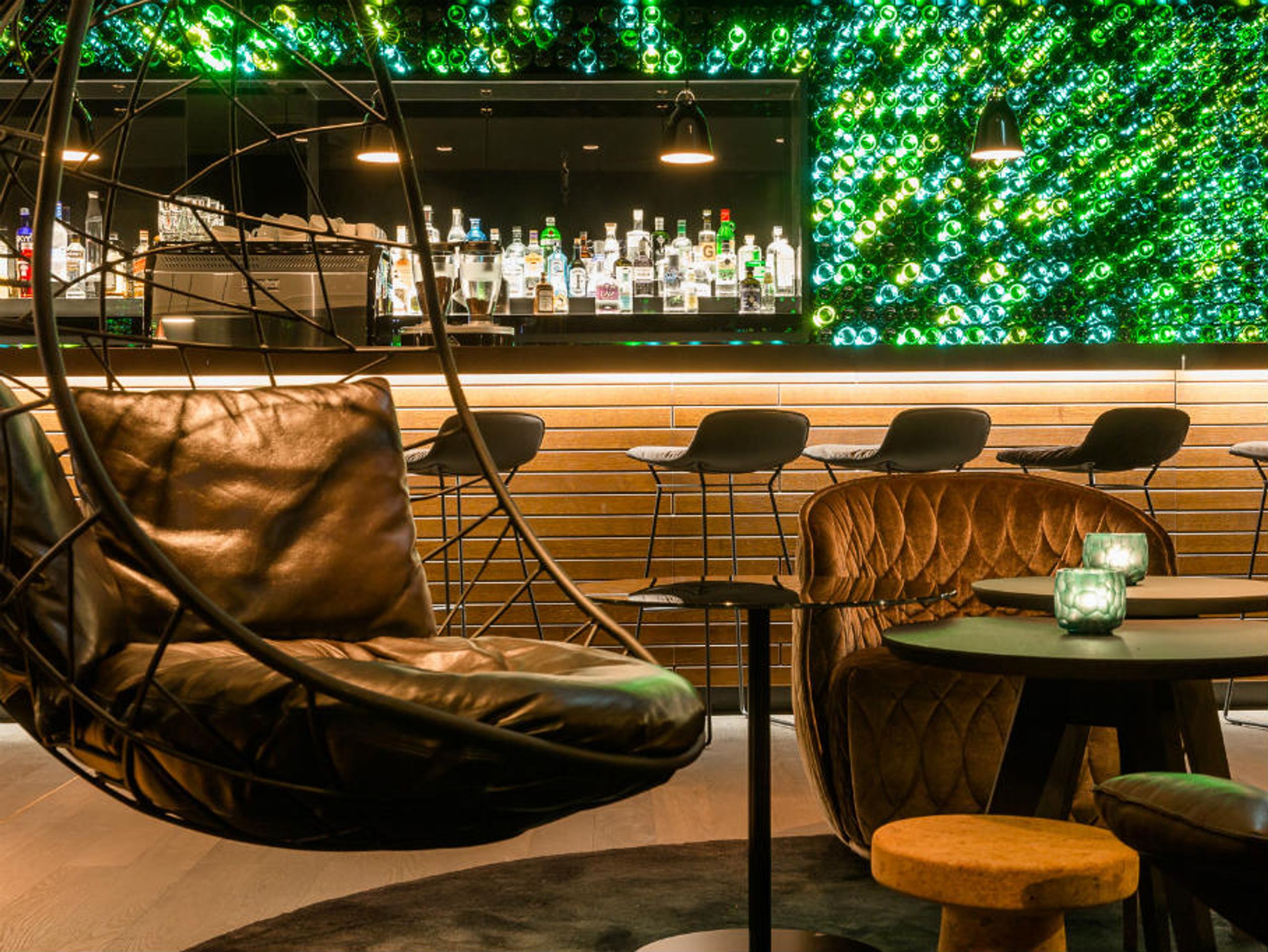 Gemütliche Hotelbar mit modernem Design, Leder-Sitzmöbeln und Hängekorb. Im Hintergrund leuchtet eine Wand aus grünen Flaschen, die für eine besondere Atmosphäre sorgt. Die Bar zeigt eine Auswahl an Spirituosen, ideal für entspannte Abende.