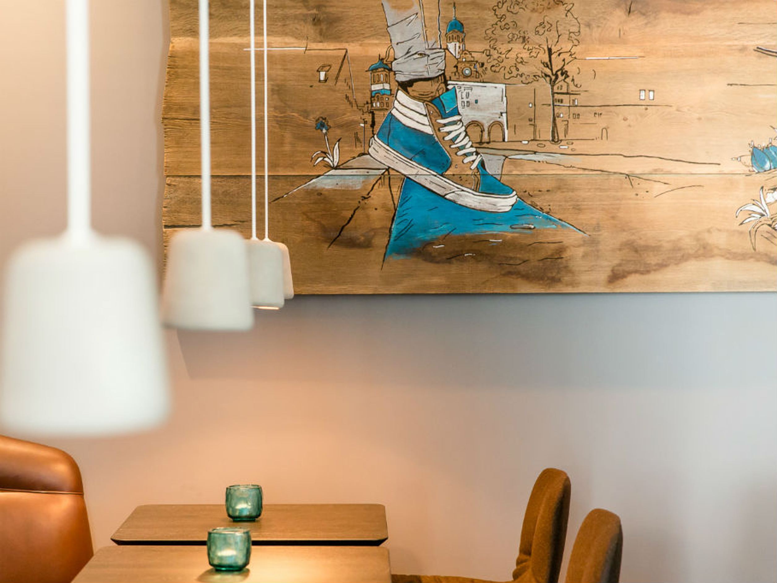 Motel One Essbereich mit zwei Tischen und Stühlen, warmes Licht durch Hängeleuchten. An der Wand hängt ein großes, farbenfrohes Gemälde mit einer blauen Turnschuhillustration.