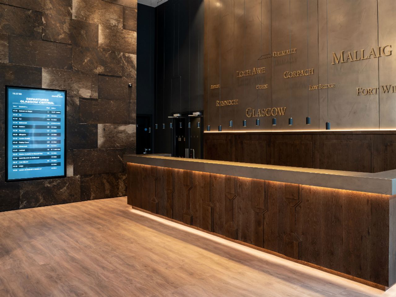 Rezeption eines modernen Hotels mit einem langen Holztresen, beleuchtetem Boden und einer digitalen Anzeigetafel an der Wand, die Informationen anzeigt. Wände sind in dunklen Farbtönen gehalten, mit eingravierten Städtenamen als dekoratives Element.