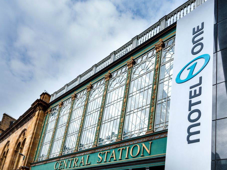 Fassade eines historischen Bahnhofsgebäudes mit großen Glasfenstern und der Aufschrift „Central Station“; daneben ein modernes Schild mit dem Logo und Namen „Motel One“. Himmel mit Wolken im Hintergrund.