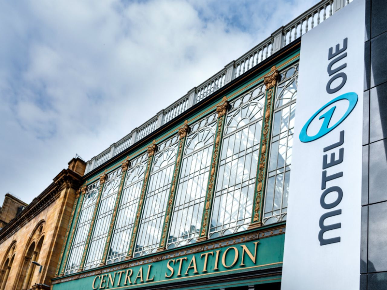 Fassade eines historischen Bahnhofsgebäudes mit großen Glasfenstern und der Aufschrift „Central Station“; daneben ein modernes Schild mit dem Logo und Namen „Motel One“. Himmel mit Wolken im Hintergrund.