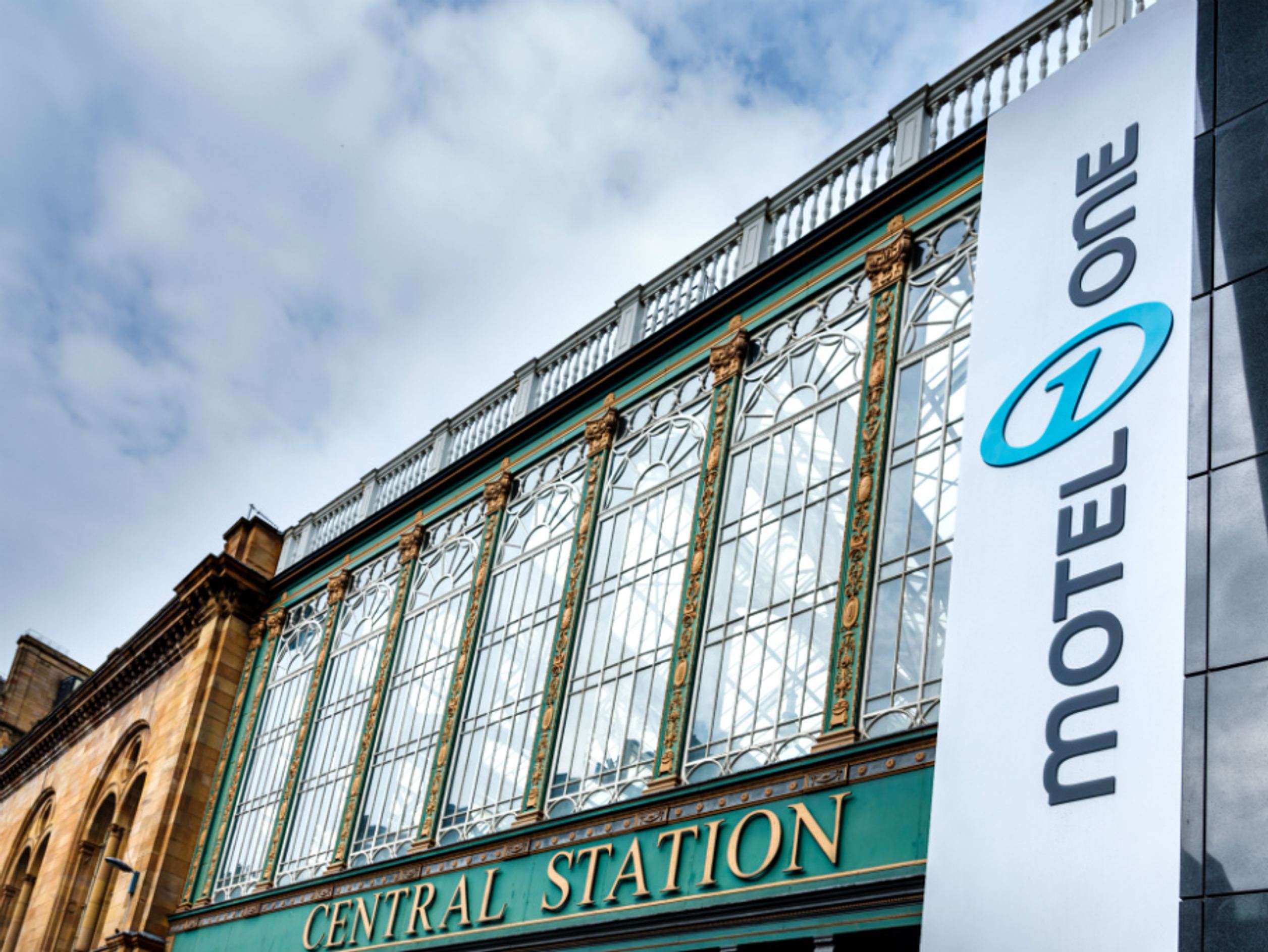 Fassade eines historischen Bahnhofsgebäudes mit großen Glasfenstern und der Aufschrift „Central Station“; daneben ein modernes Schild mit dem Logo und Namen „Motel One“. Himmel mit Wolken im Hintergrund.