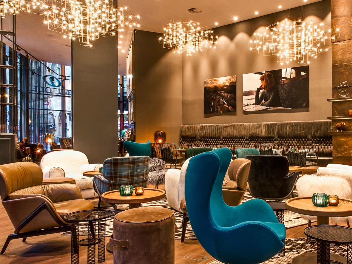 Motel One Hotel-Lounge mit bequemen Sesseln und Sofas in verschiedenen Farben, stimmungsvoller Beleuchtung durch dekorative Deckenleuchten und kleinen Tischen mit Kerzen