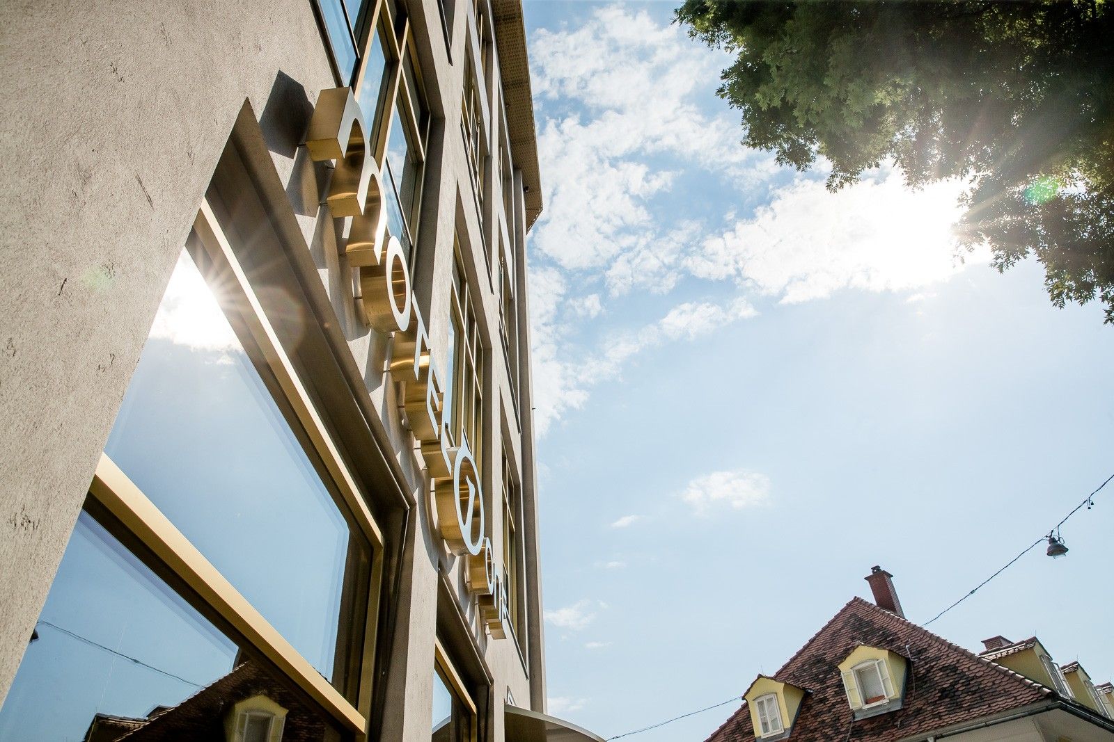 Blick nach oben auf die moderne Fassade eines Hotels mit großem, reflektierendem Fenster, darunter die Hotelbeschriftung. Im Hintergrund sind Dachkuppeln traditioneller Häuser und ein sonniger Himmel mit wenigen Wolken zu sehen.